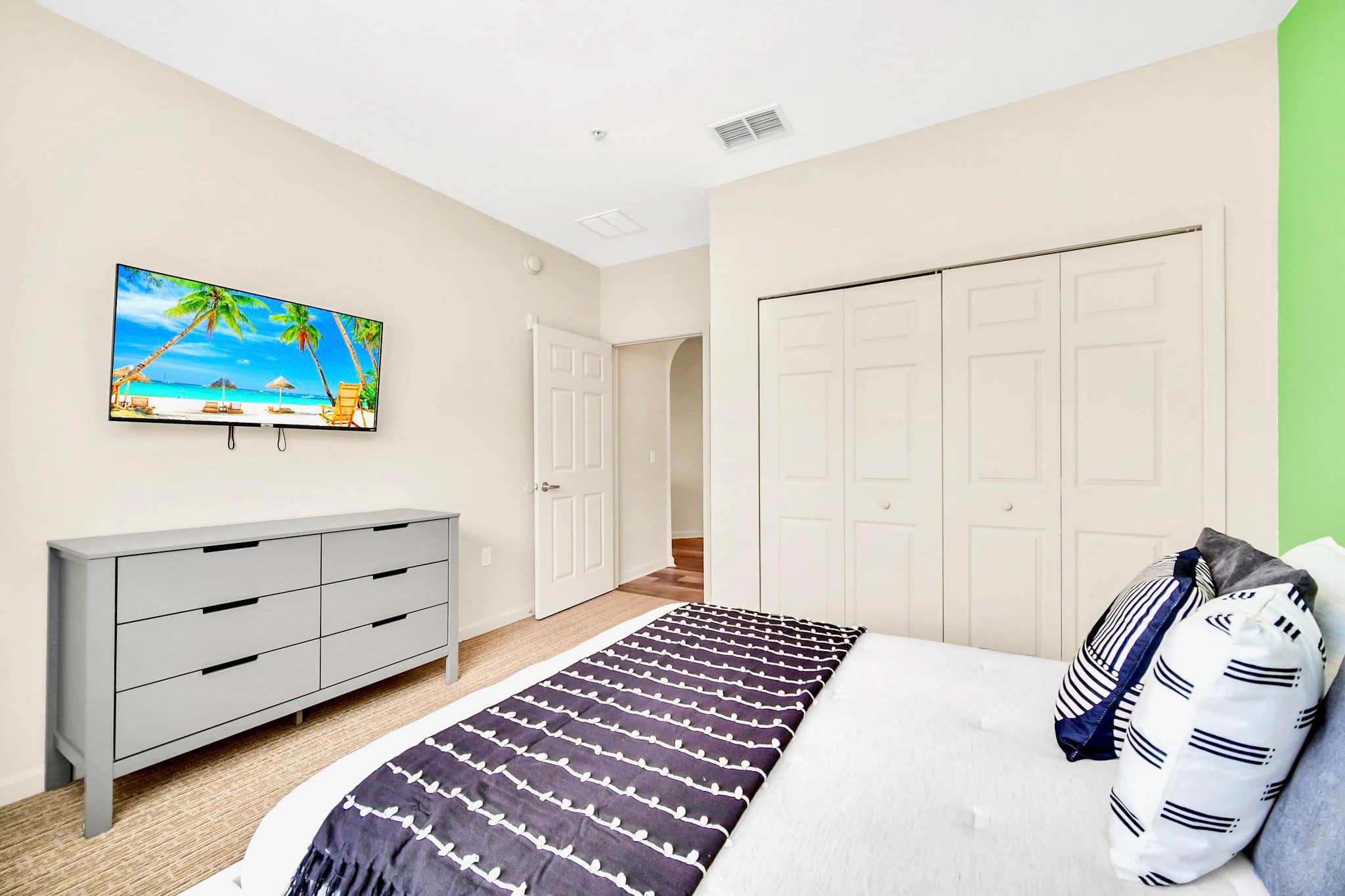 View of the second bedroom showing a white platform bed with blue and white bedding, a grey dresser, and a wall-mounted TV. The room has a green accent wall and bi-fold closet doors.