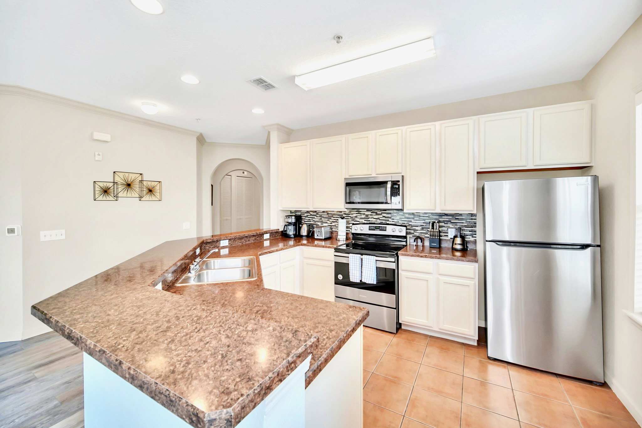 Well-equipped kitchen featuring light cream cabinetry, brown speckled countertops, a stainless steel refrigerator, microwave, and electric range, with a mosaic tile backsplash.