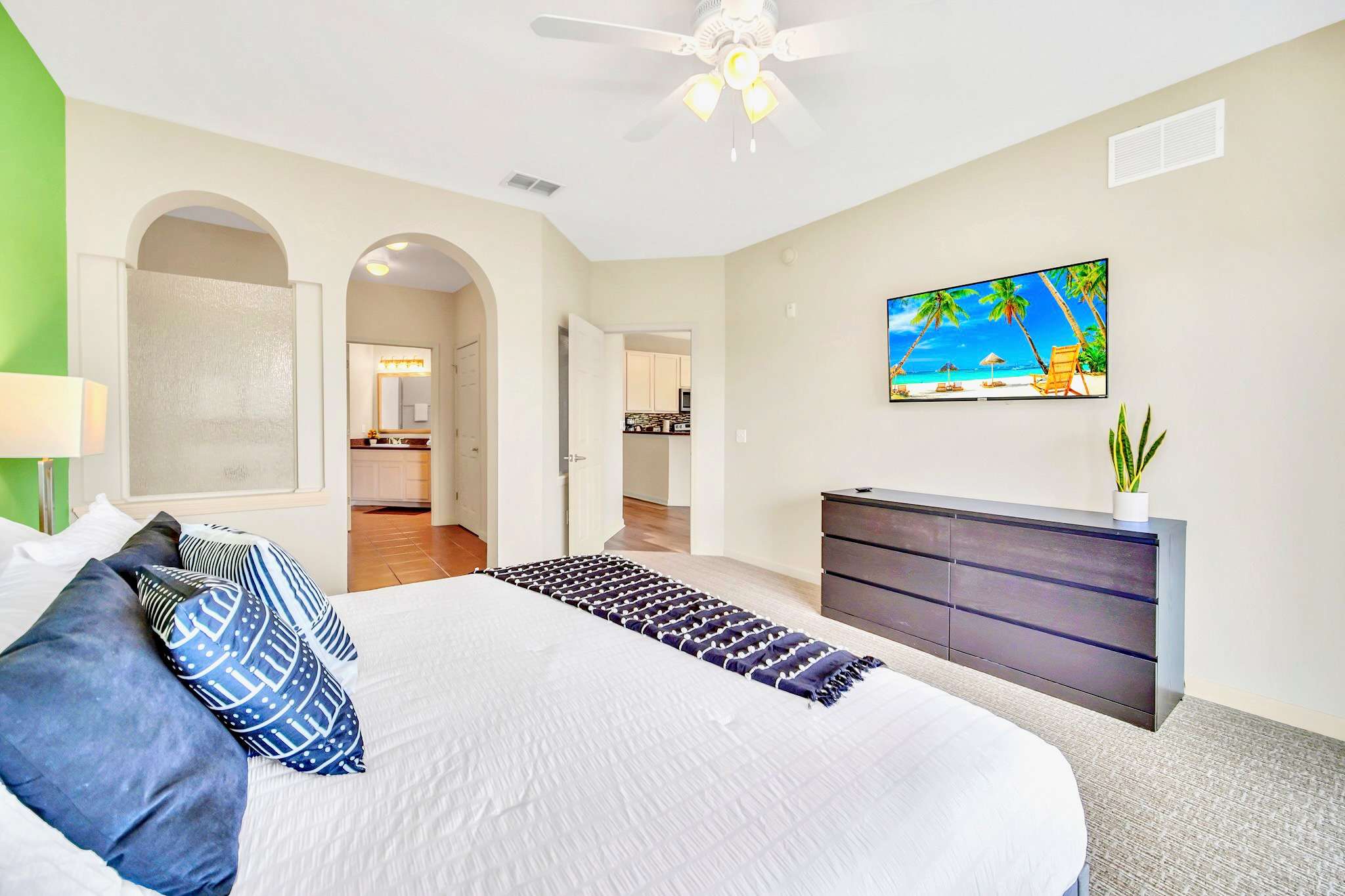 Spacious master bedroom with a large bed featuring blue and white patterned pillows and a dark throw, a dark wood dresser, and a wall-mounted TV displaying a beach scene. The room has light grey carpet and a ceiling fan, with arched doorways leading to a bathroom.
