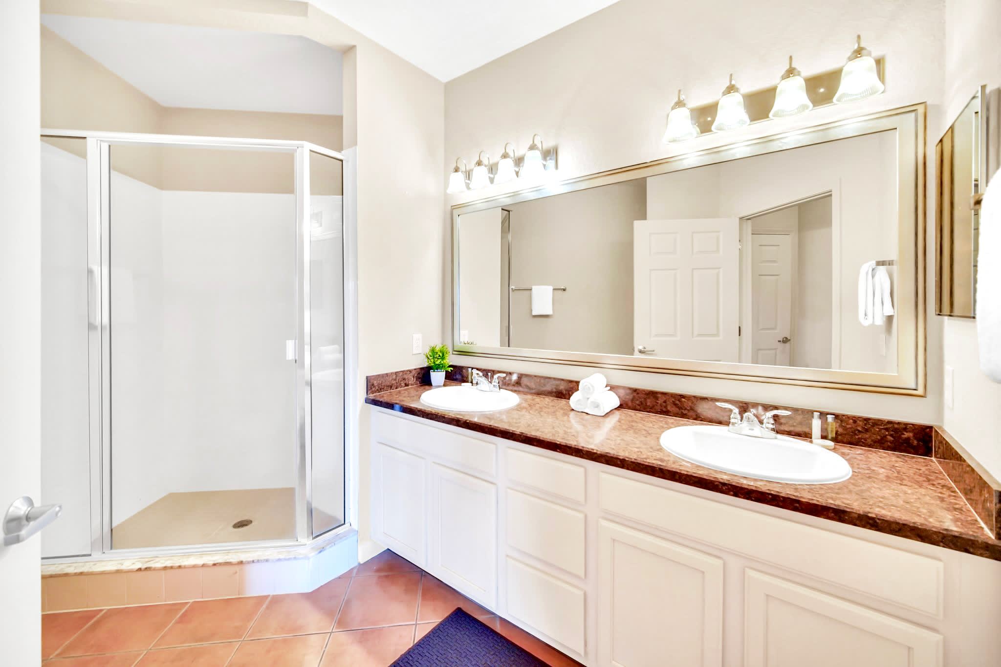 Master bathroom with a double vanity featuring white cabinets and dark brown granite countertops, a large mirror, and a glass-enclosed walk-in shower. The floor is covered in terracotta-colored tiles.