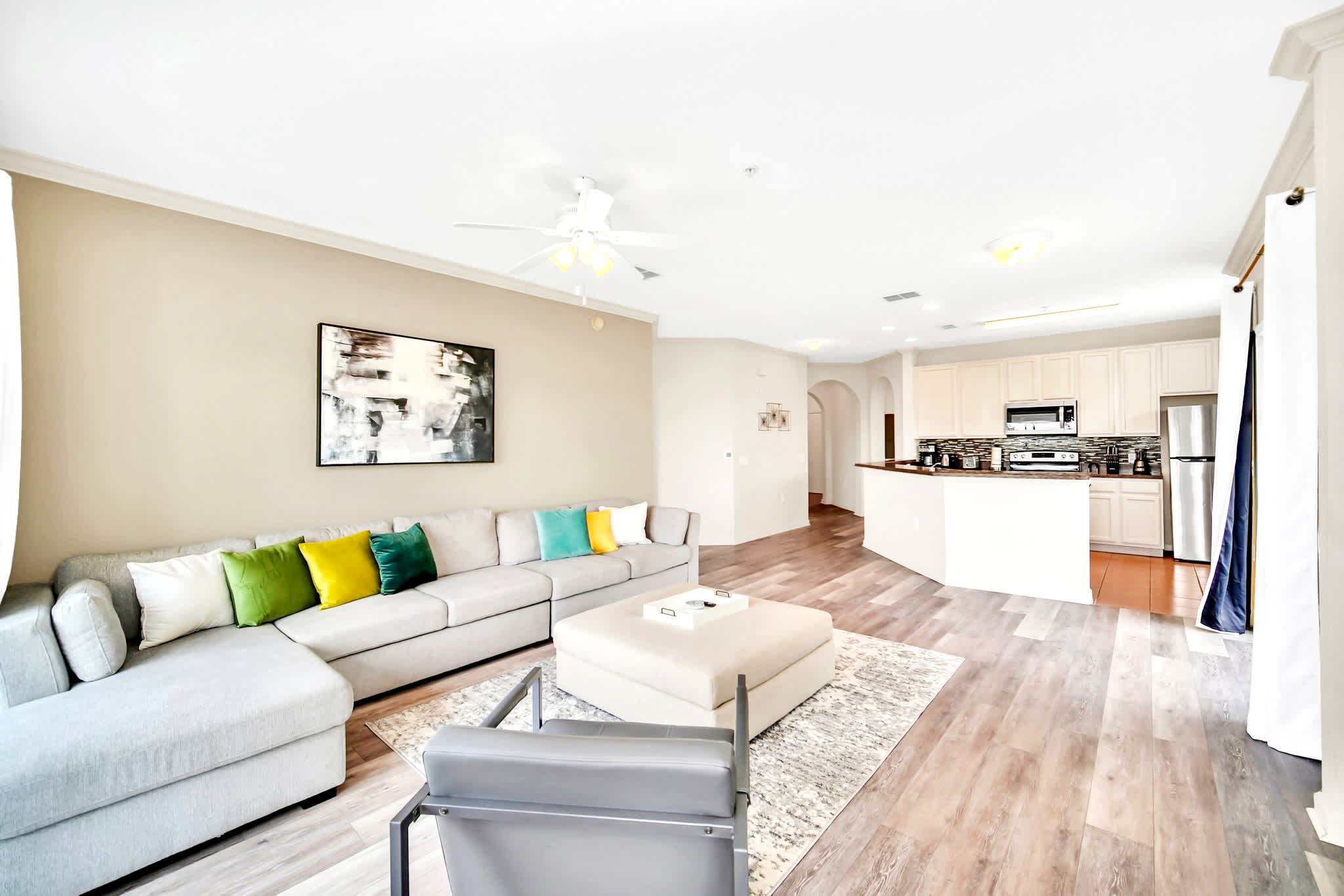 Open-concept living space with a large L-shaped sectional sofa, ottoman, and wood-look flooring, flowing into a kitchen with light cabinetry and a breakfast bar, all under a ceiling fan.