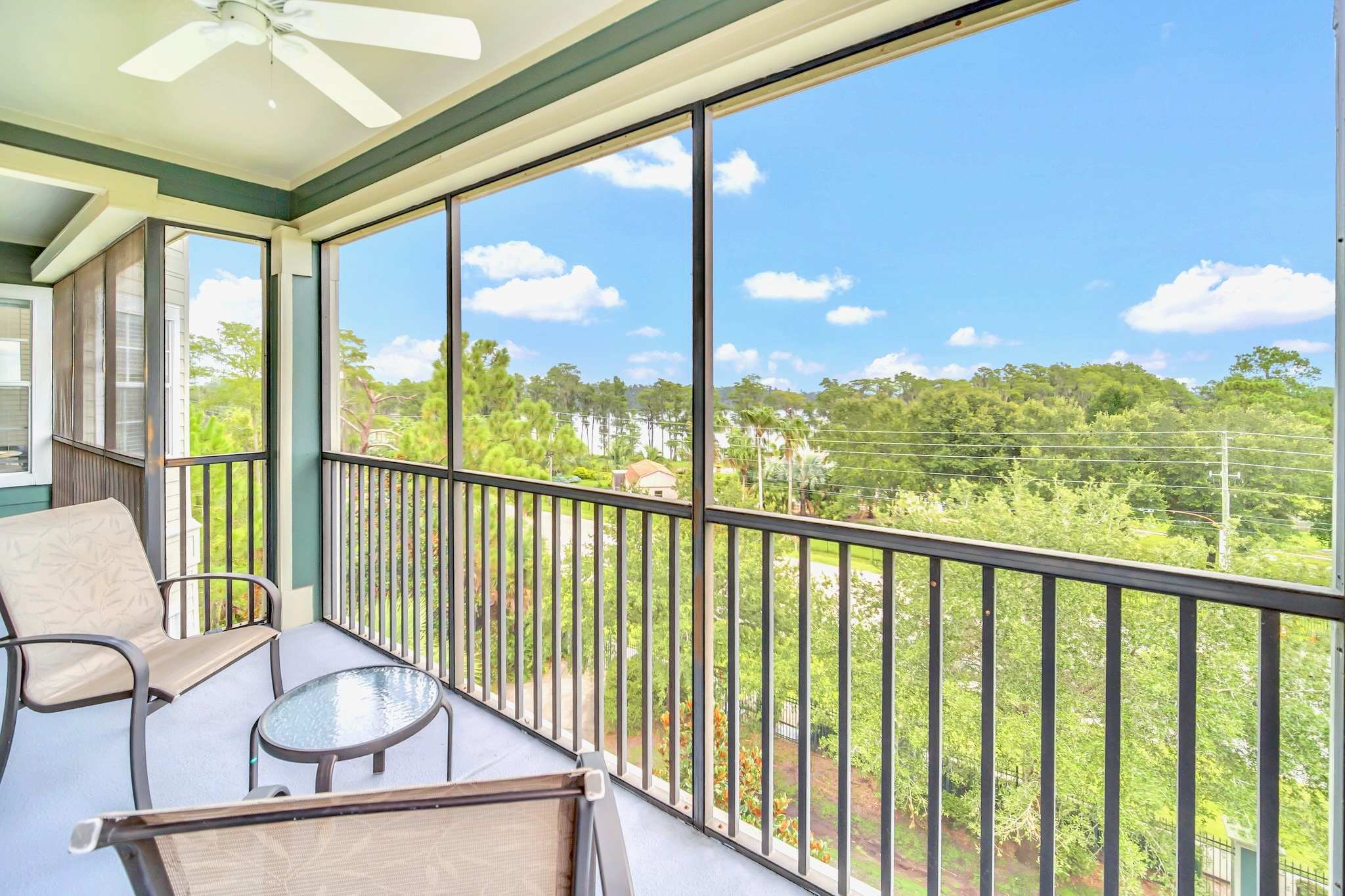 A screened-in balcony with outdoor seating, offering a view of lush green trees and a clear blue sky. The balcony features a ceiling fan and black metal railings.