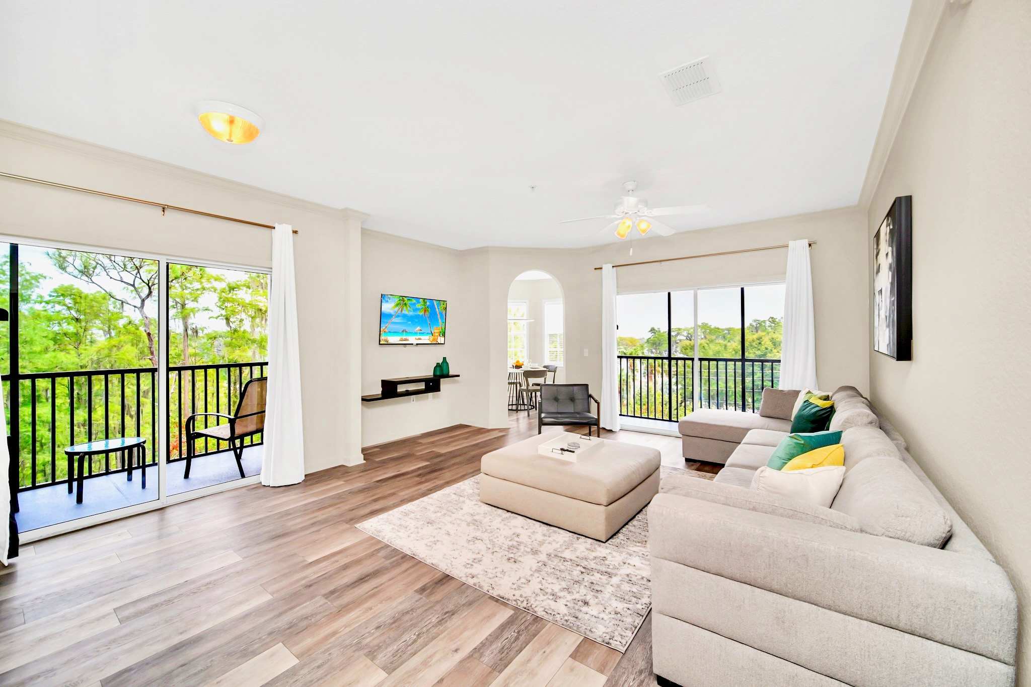 Inviting living room with light wood-look flooring, a large L-shaped sectional sofa, a beige ottoman, and a wall-mounted TV, with a wide sliding glass door opening to a screened balcony.