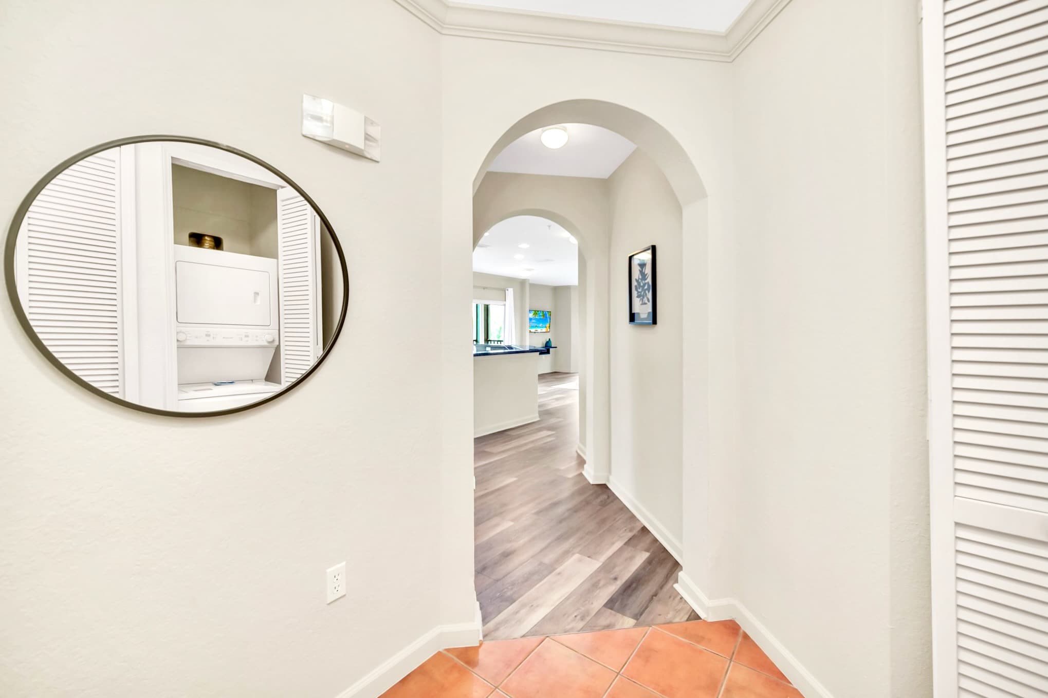 A bright hallway with terracotta-colored tile flooring transitioning to wood-look flooring, featuring arched doorways and a round mirror reflecting a stacked washer and dryer unit.