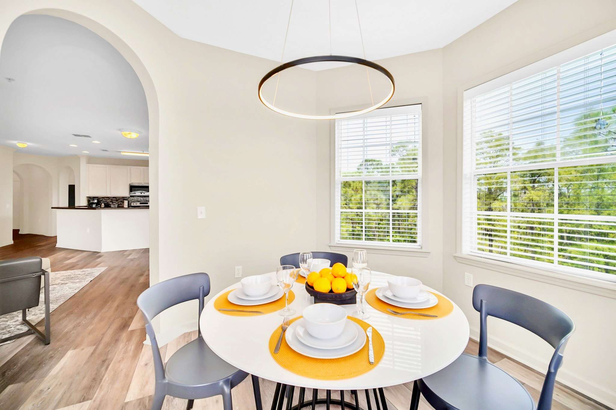 Bright dining area with a round white table and gray chairs, featuring a modern circular pendant light and bay windows, with a glimpse of the kitchen's breakfast bar in the background.