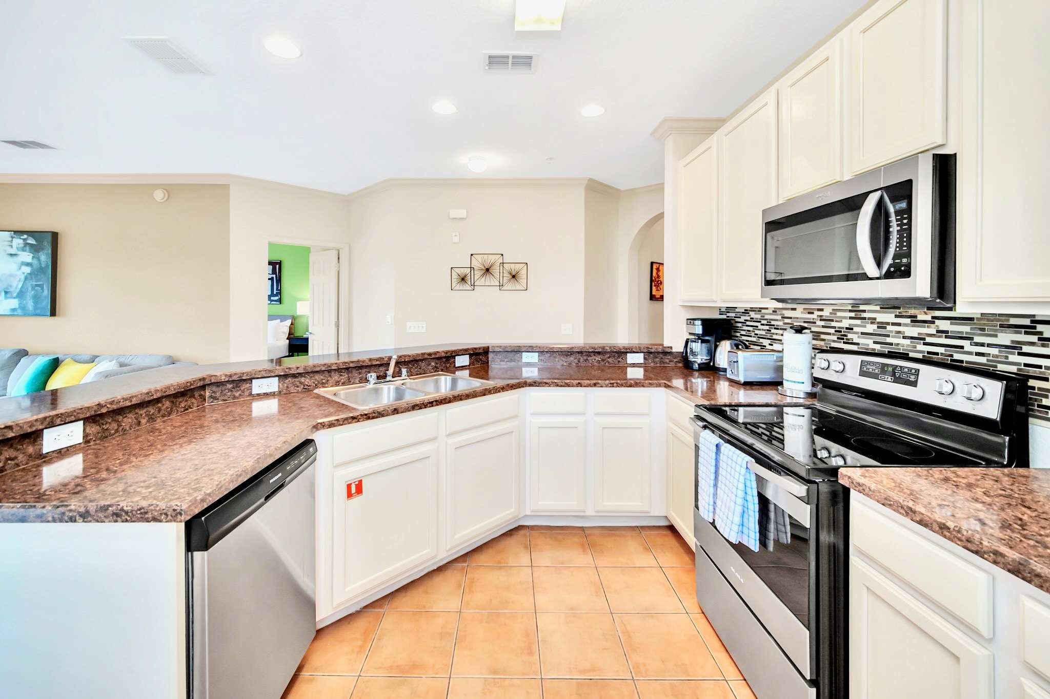 A modern kitchen featuring light cream cabinetry, dark brown granite countertops, and a mosaic tile backsplash. Stainless steel appliances include a microwave, oven, and dishwasher, with terracotta-colored tile flooring.
