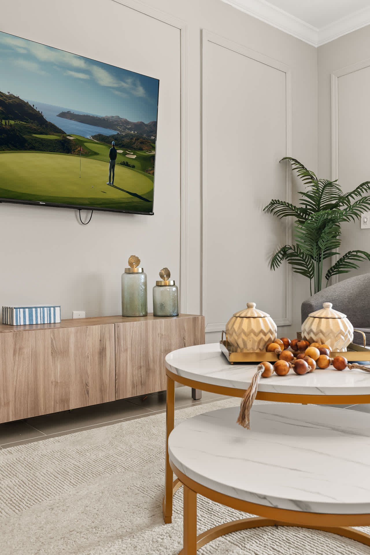 A close-up of the living room's modern decor, showcasing a nesting marble-top coffee table with decorative items, a wooden TV console, and a large television displaying a golf course scene.