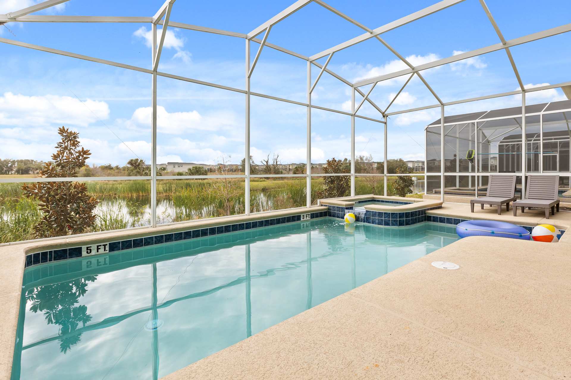 Screened-in private pool area with a hot tub, overlooking a serene lake and natural landscape under a bright blue sky. The pool deck has lounge chairs and pool floats, offering a relaxing outdoor space.
