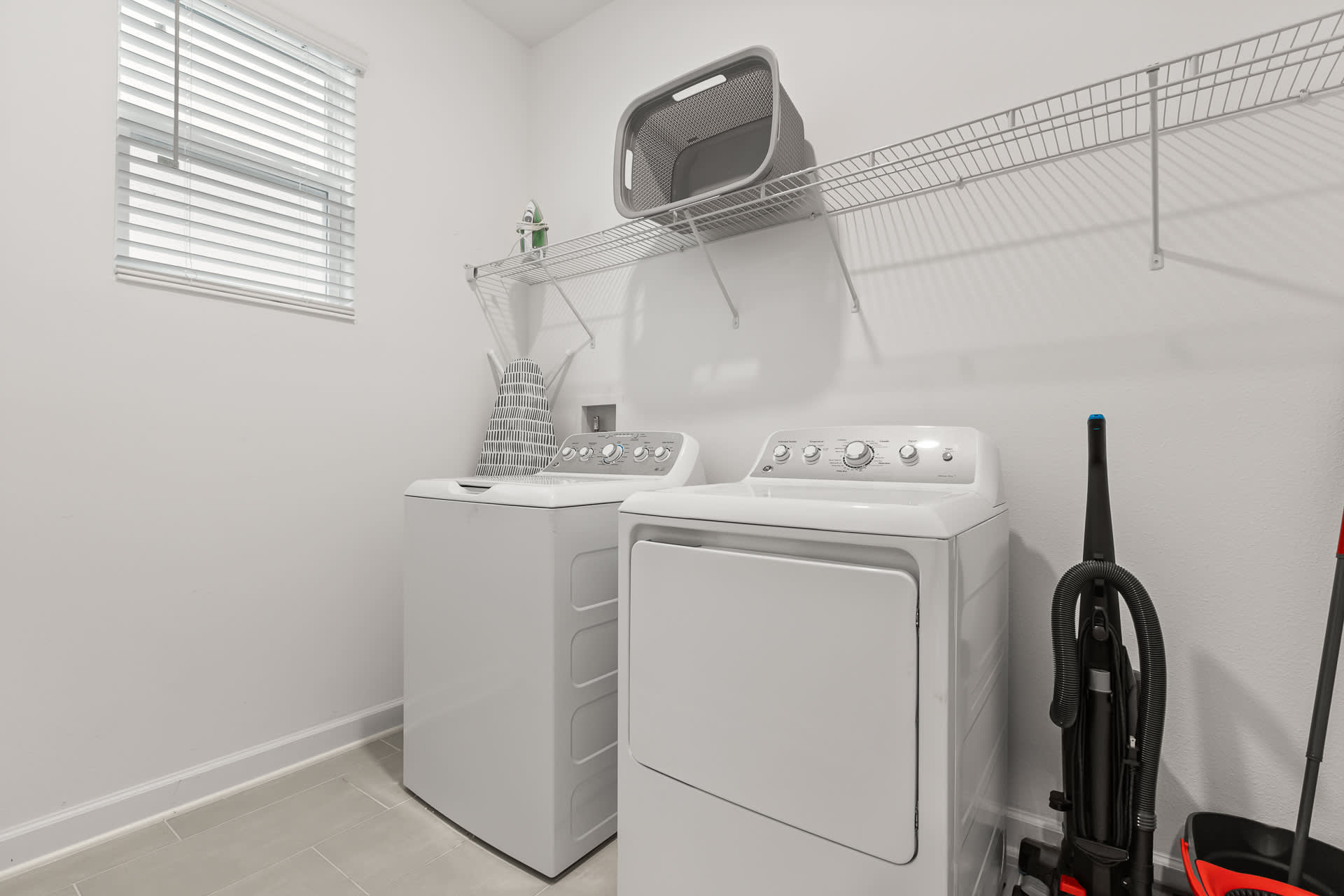 A functional laundry room equipped with a white top-loading washing machine and a front-loading dryer. A wire shelf above holds laundry essentials, and a window with blinds provides natural light.