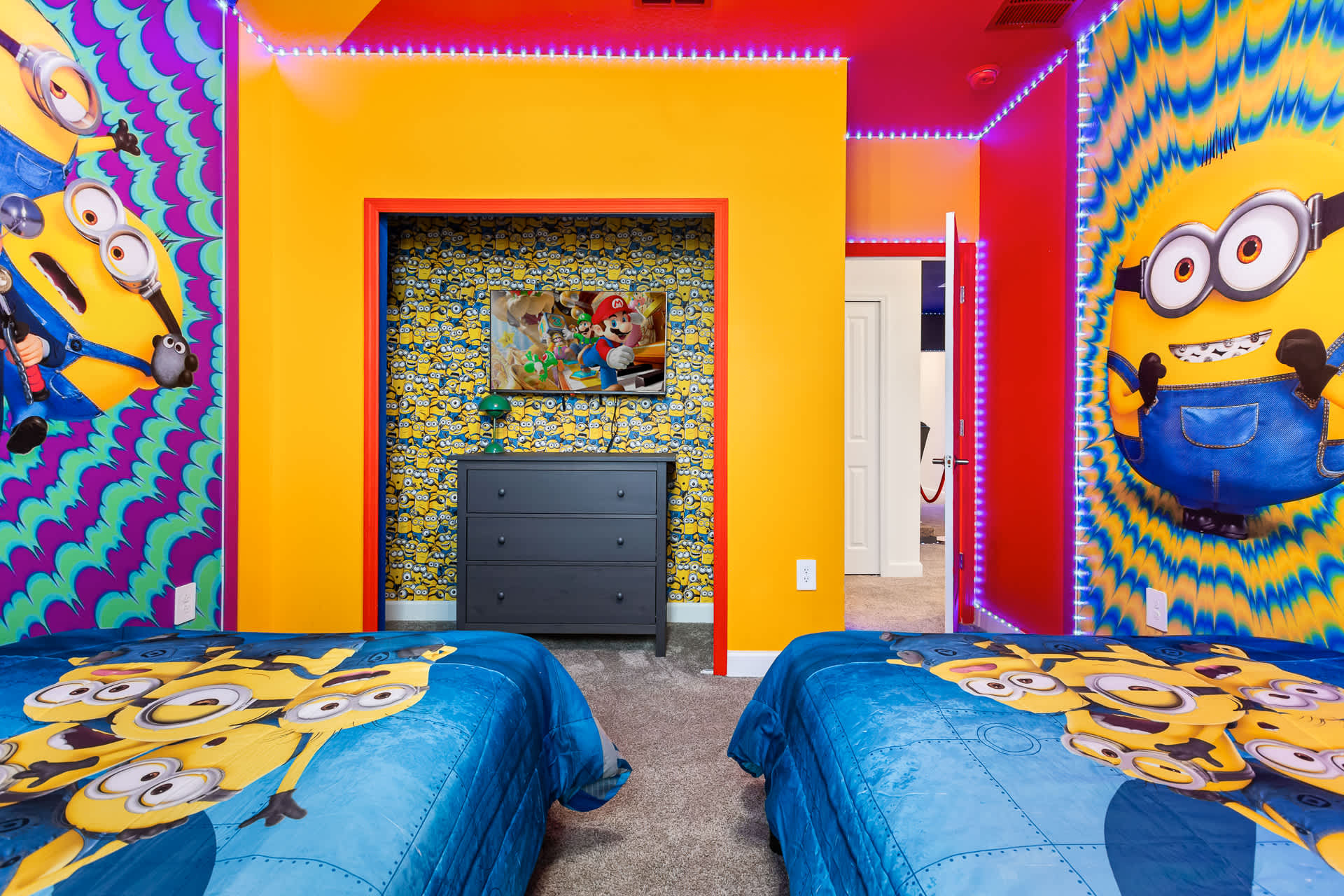 A vibrant Minions-themed bedroom featuring two twin beds with blue Minions bedding. The walls are covered in bright yellow and blue Minions wallpaper, with red LED strip lighting along the ceiling and a TV recessed into a yellow wall niche.