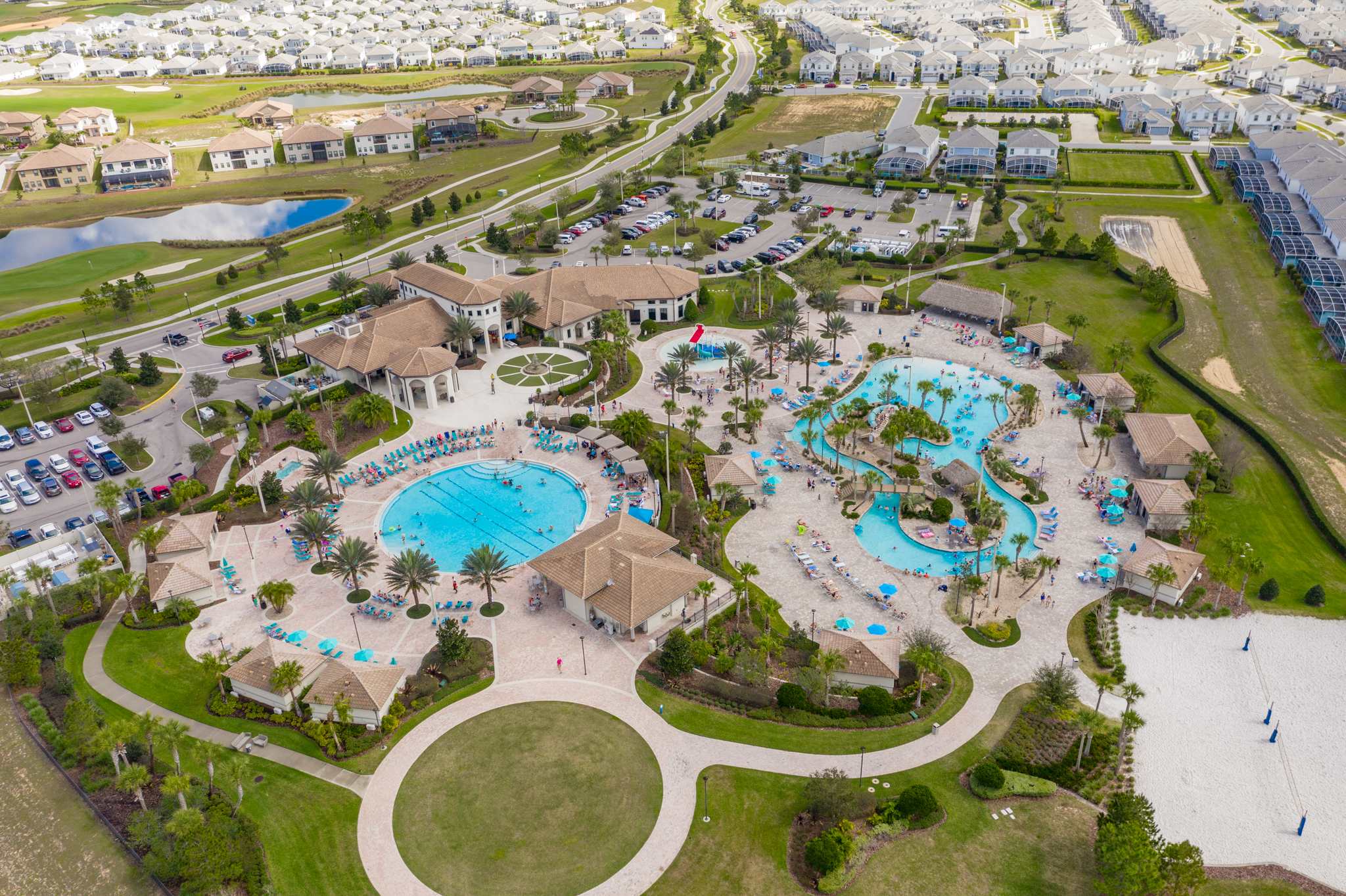 Aerial view of a large, luxurious communal resort pool area with multiple swimming pools, palm trees, lounge chairs, and surrounding buildings. The complex is set within a residential community, indicating shared amenities.