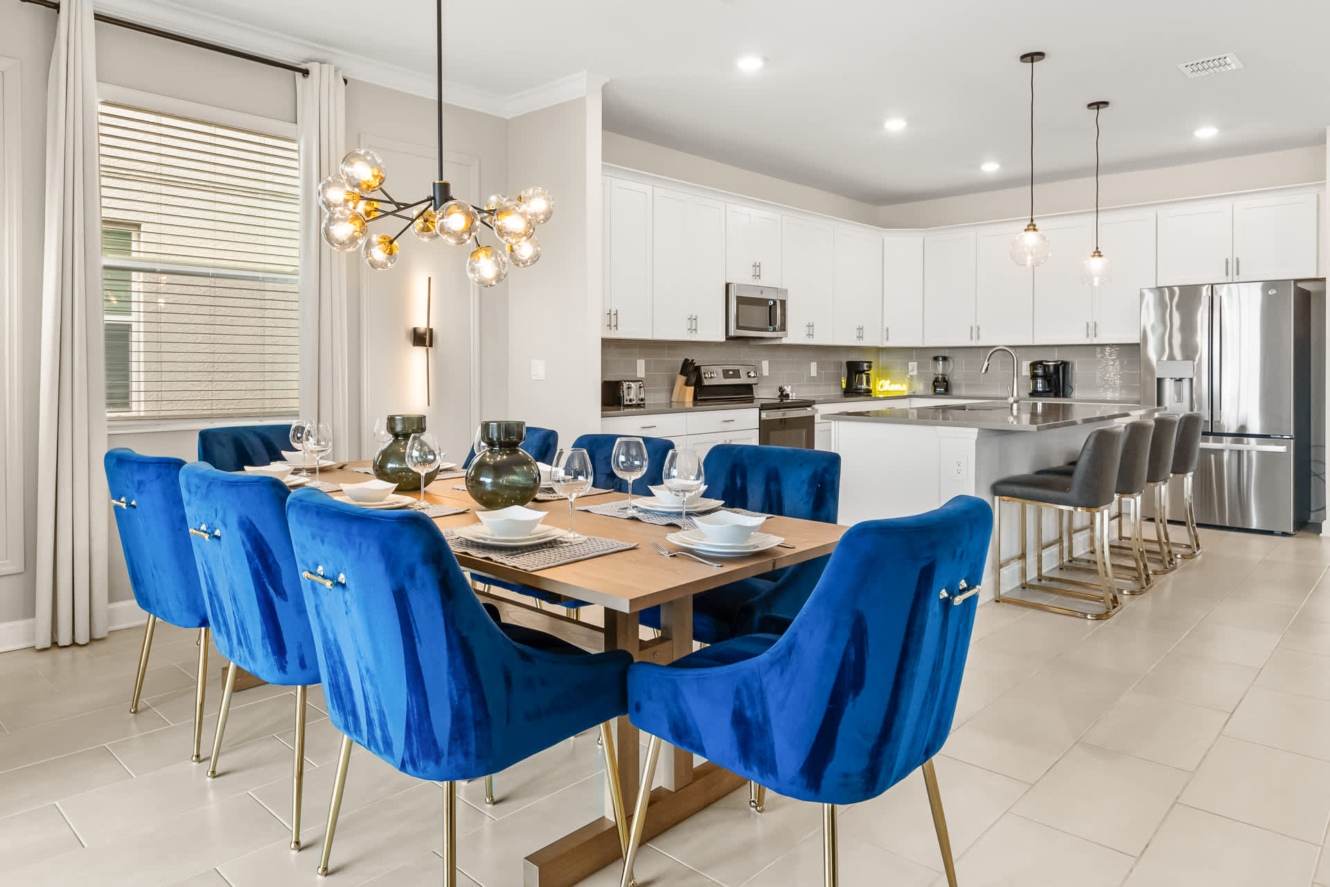 An open-concept dining area and kitchen, featuring a wooden dining table with blue velvet chairs, a modern chandelier, and a contemporary kitchen with white cabinetry, stainless steel appliances, and a large island with bar stools.
