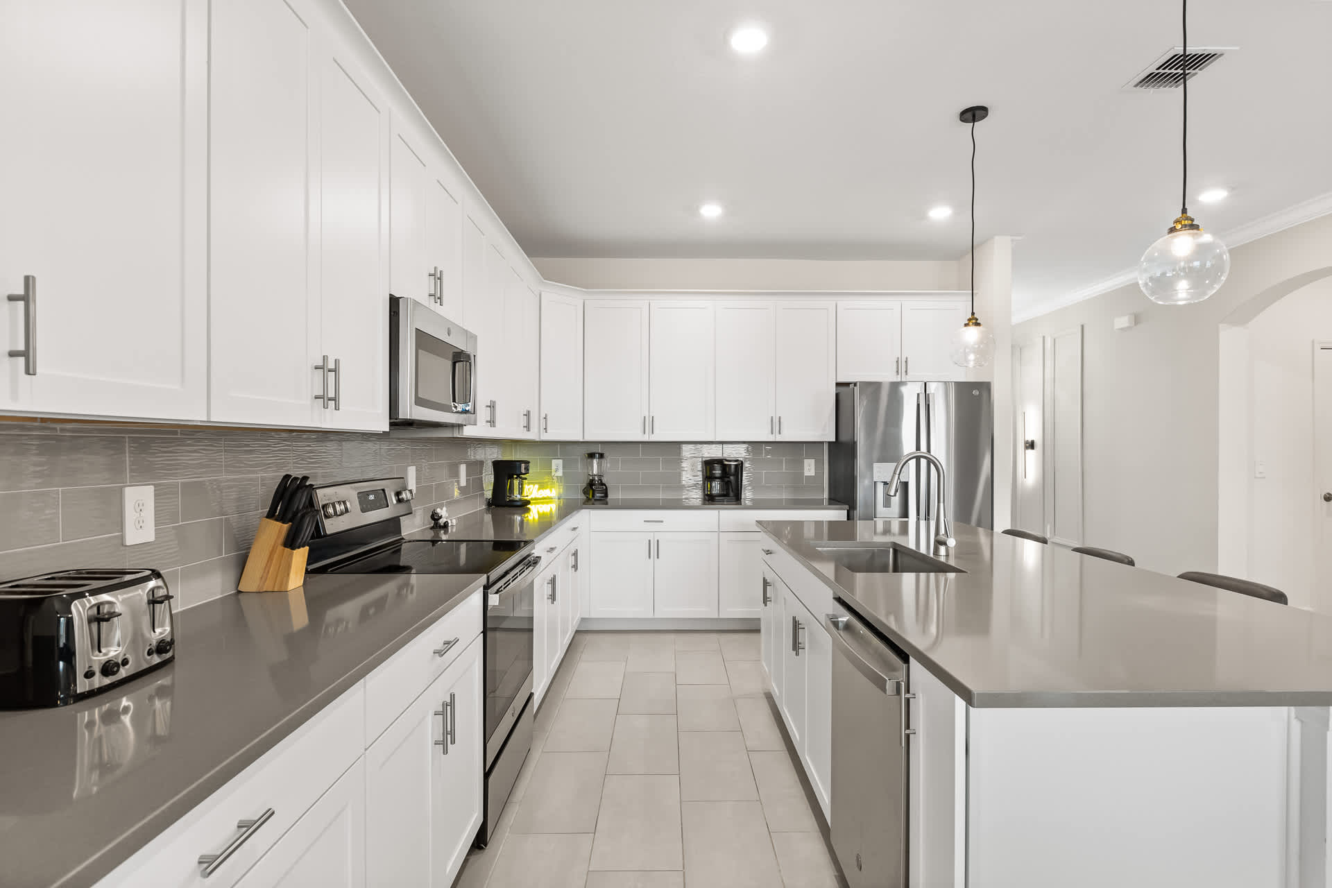 A wide view of the modern kitchen, showcasing white shaker cabinets, gray quartz countertops, stainless steel appliances, and a gray subway tile backsplash, with a large island and a neon "Hello" sign.