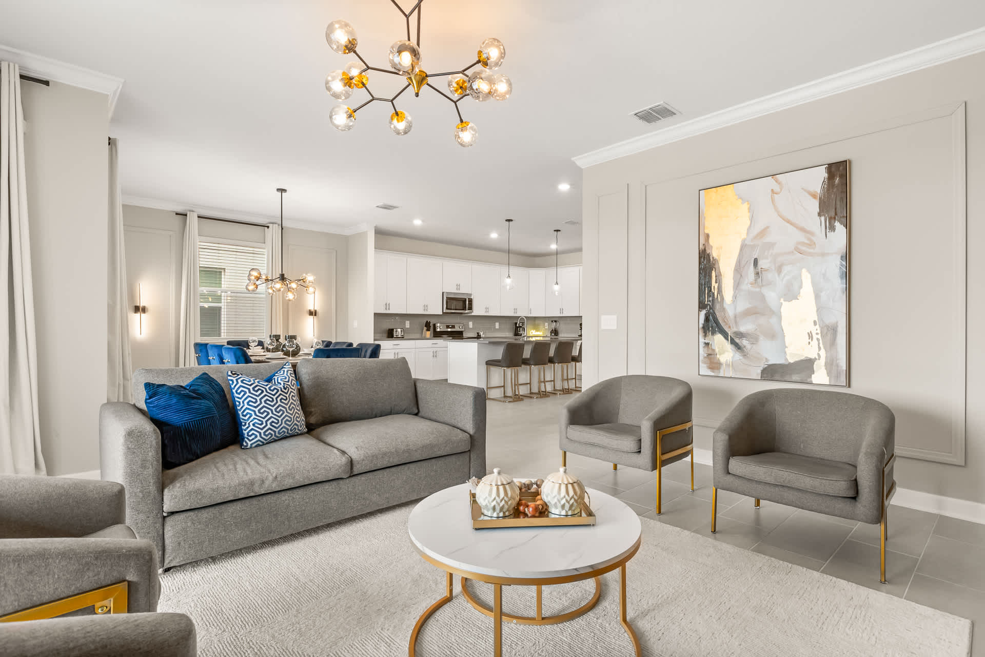 Modern open-concept living room with light gray walls and tile flooring, furnished with a gray sofa and two gray armchairs around a round coffee table. A stylish gold chandelier and abstract art piece add elegance to the space, which flows into the dining and kitchen areas.