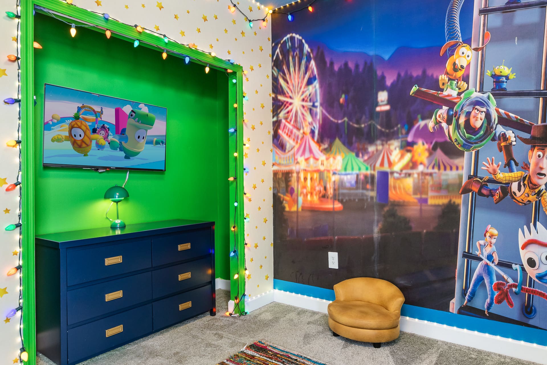 A corner of the themed kids' bedroom, featuring a vibrant green accent wall with a wall-mounted TV above a blue dresser, surrounded by string lights and part of the Toy Story mural.