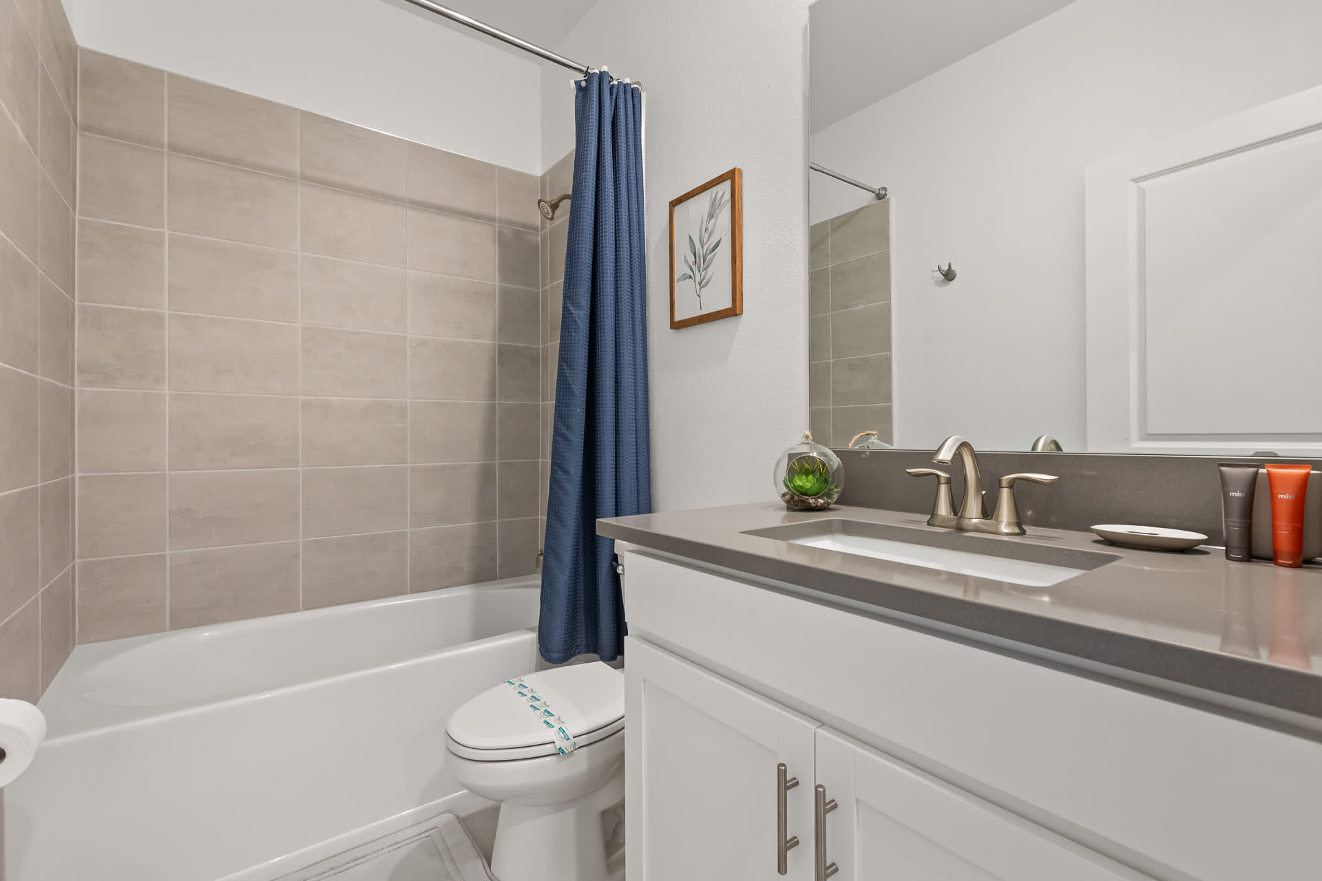 A clean bathroom featuring a white vanity with a grey countertop and a rectangular sink, alongside a toilet and a shower-tub combo with light grey tiled walls. A blue shower curtain adds a pop of color, and a framed botanical print hangs on the wall.