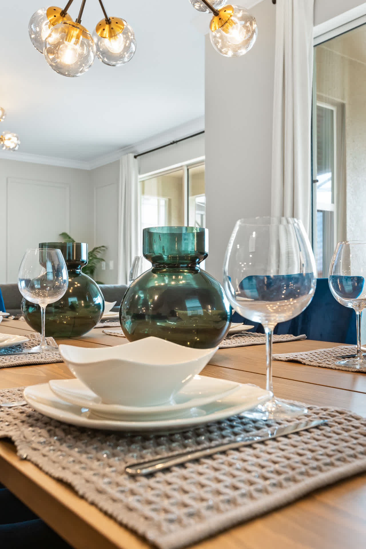 A detailed view of the dining table set with white dinnerware, wine glasses, and two large green glass vases, highlighting the textured placemats and the modern chandelier above.