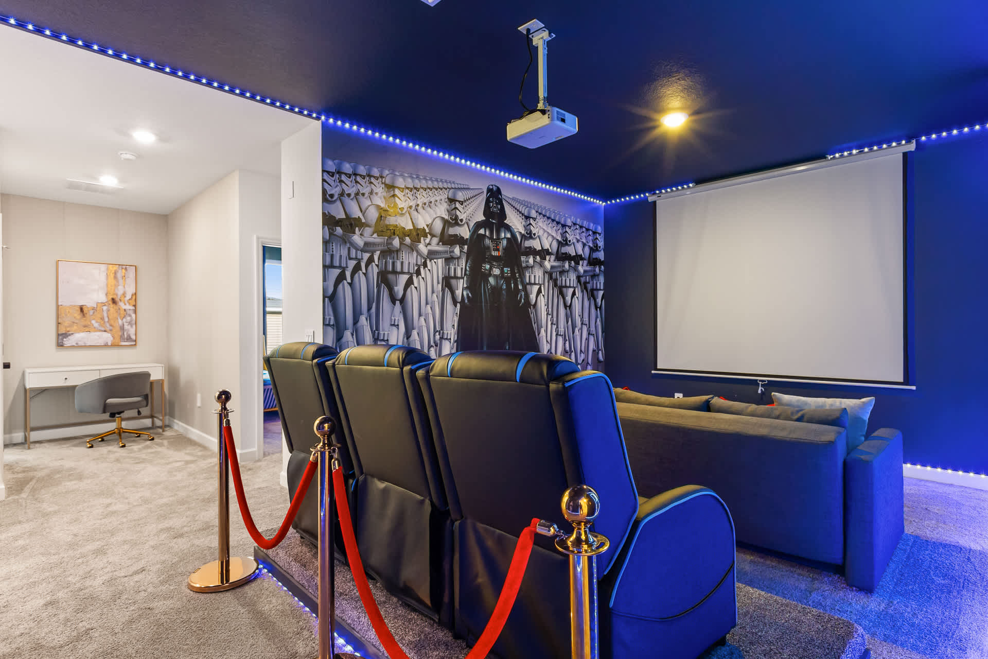 Themed home cinema room with dark blue walls and ceiling, featuring a large projection screen and comfortable reclining seats. A dramatic Darth Vader and Stormtrooper mural covers one wall, enhanced by blue LED strip lighting.