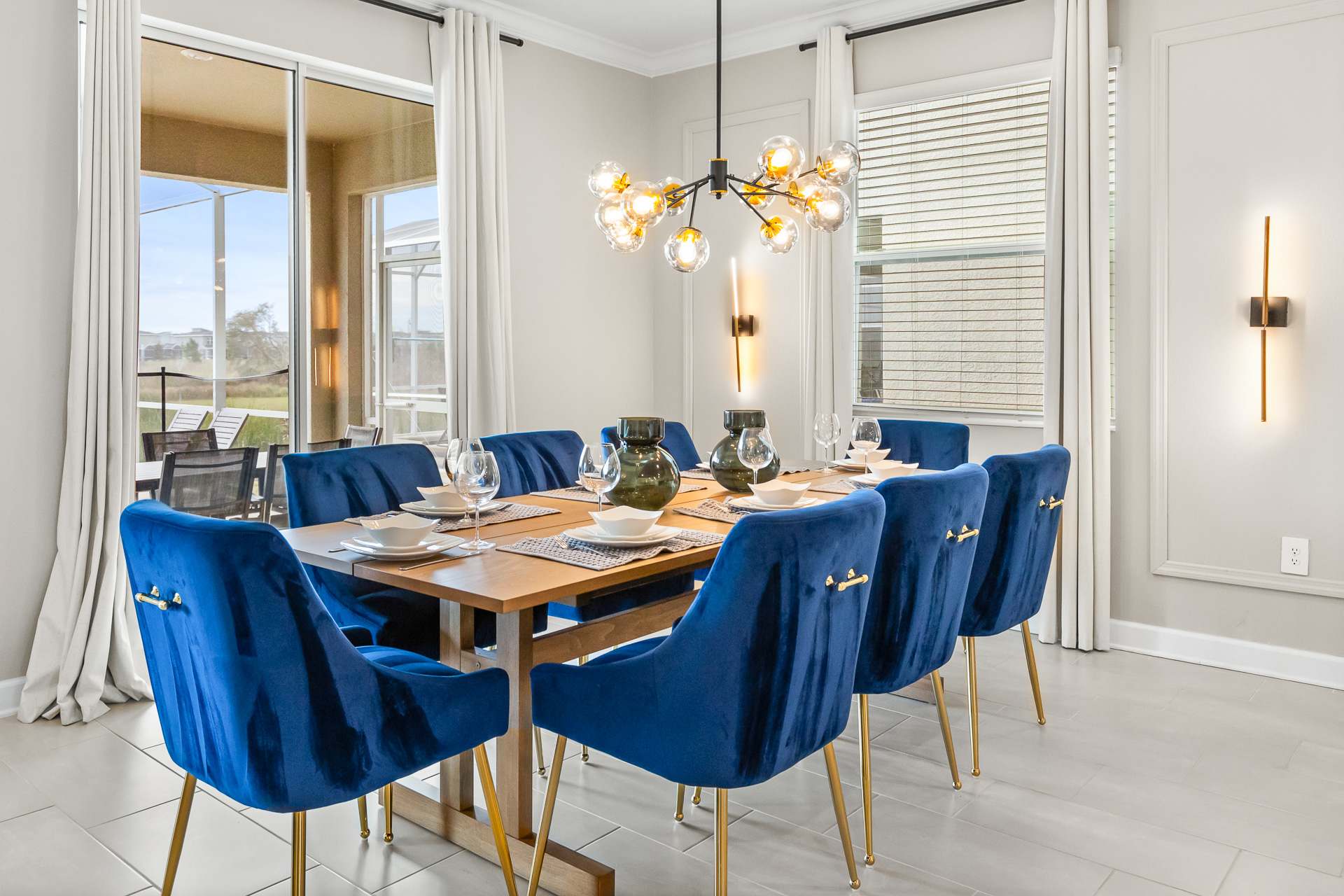 A chic dining room featuring a long wooden dining table surrounded by eight royal blue velvet chairs with gold legs, illuminated by a modern Sputnik-style chandelier, with large windows providing natural light.