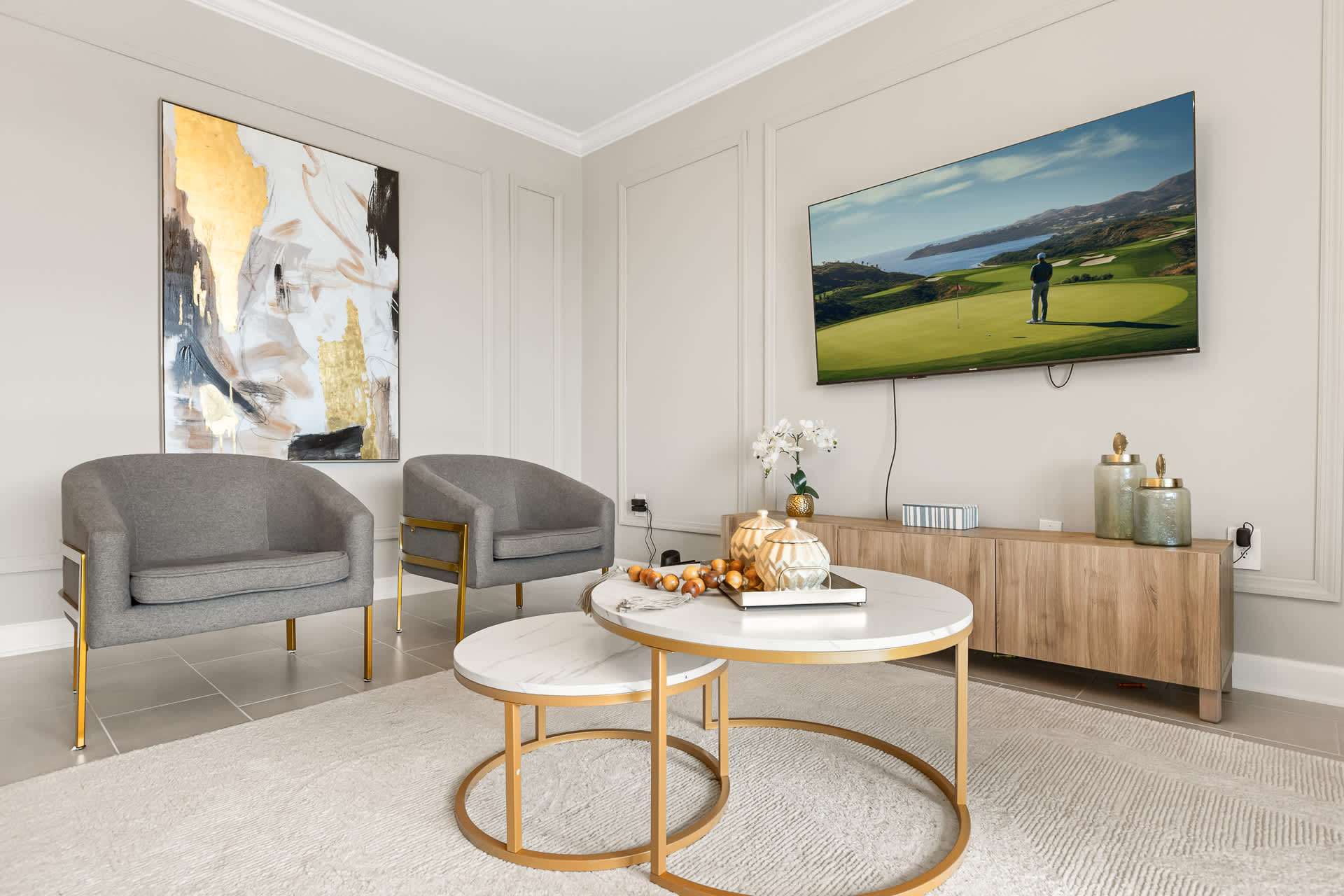 A stylish living room featuring two gray armchairs, a nesting marble-top coffee table, and a large abstract gold and gray painting, with a television mounted on the light gray paneled wall.