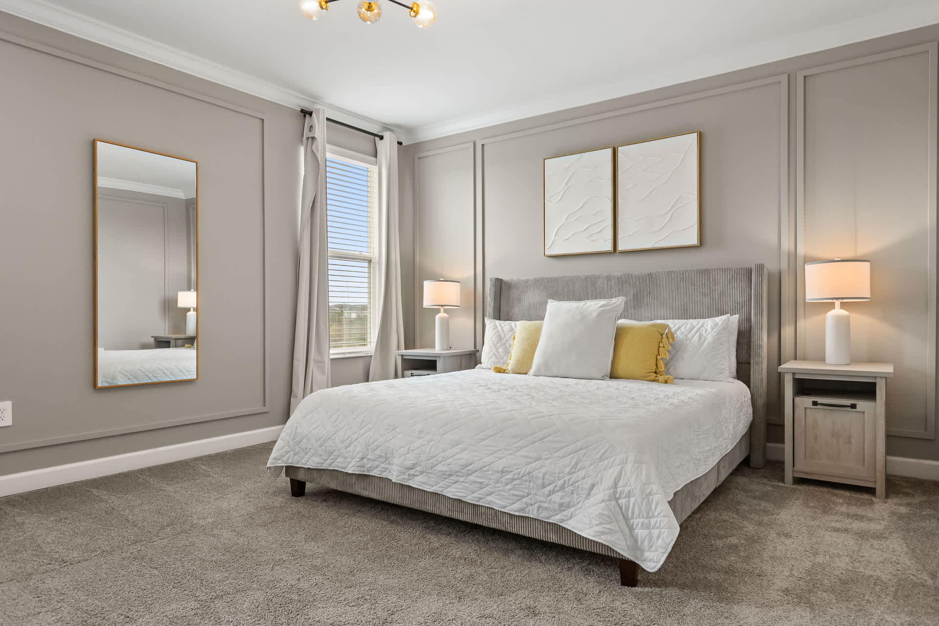 Spacious bedroom with a queen-sized bed, gray upholstered headboard, light gray carpet, and light gray paneled walls, featuring two nightstands with lamps and a full-length mirror.
