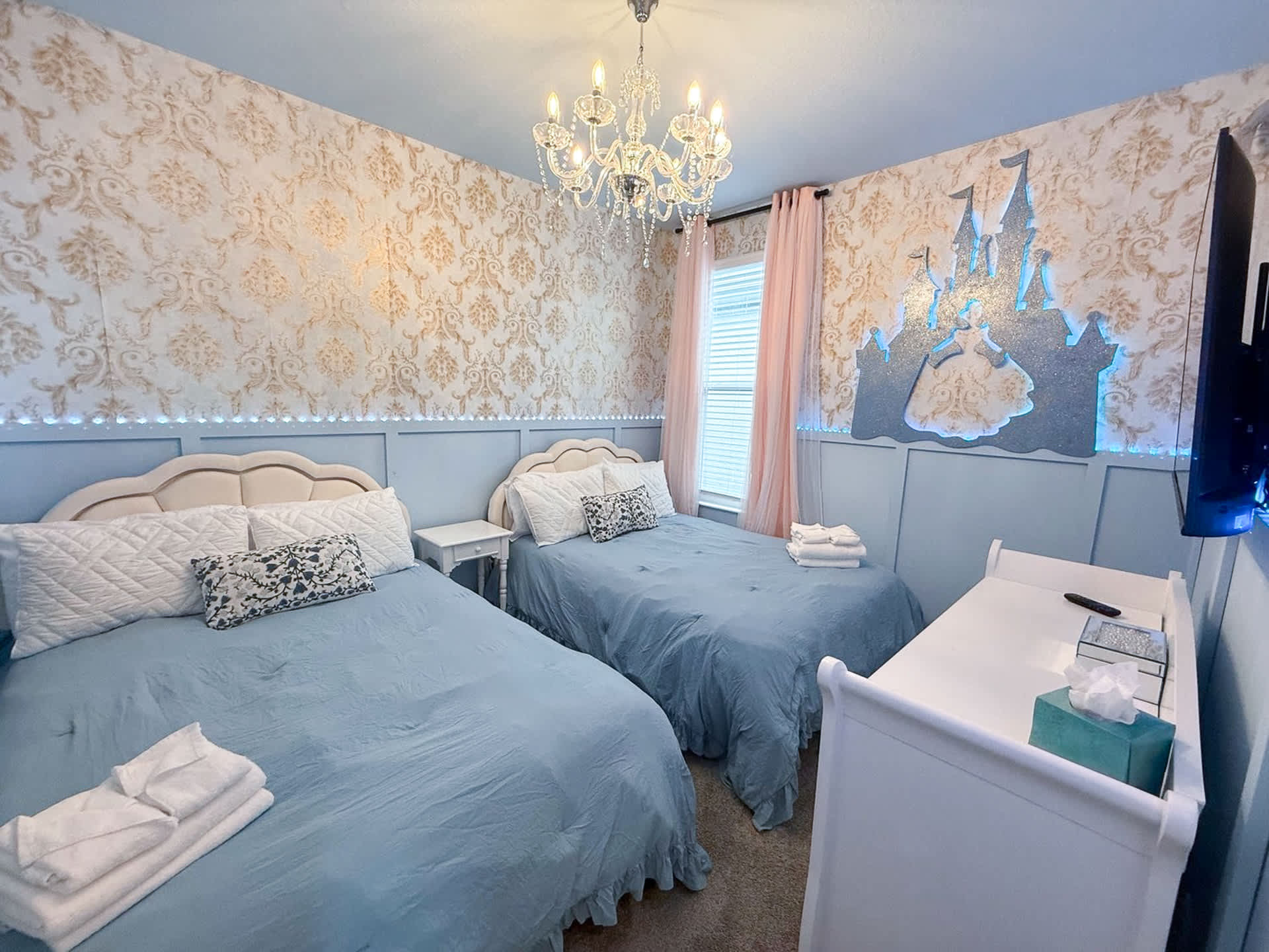 A princess-themed bedroom with two twin beds featuring light blue bedding and white headboards. The walls are adorned with ornate gold and white wallpaper above light blue wainscoting, and a large Cinderella's castle cutout with LED lighting decorates one wall.