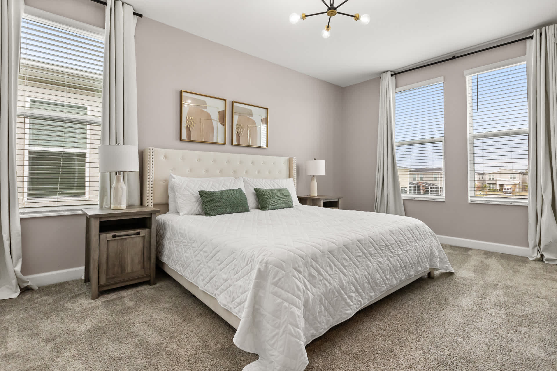 A spacious bedroom featuring a white tufted headboard king-sized bed with white quilted bedding and two green accent pillows. Two dark wood nightstands with white lamps flank the bed, and large windows with blinds and light curtains provide natural light.