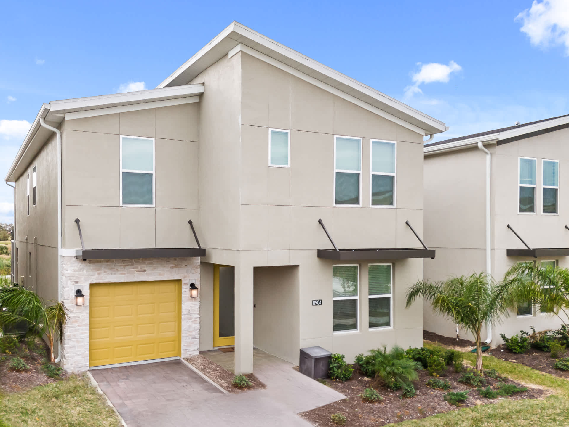 Modern two-story house exterior with light beige stucco siding, multiple windows, and a bright yellow garage door and front door. The property features a paved driveway and minimal landscaping.