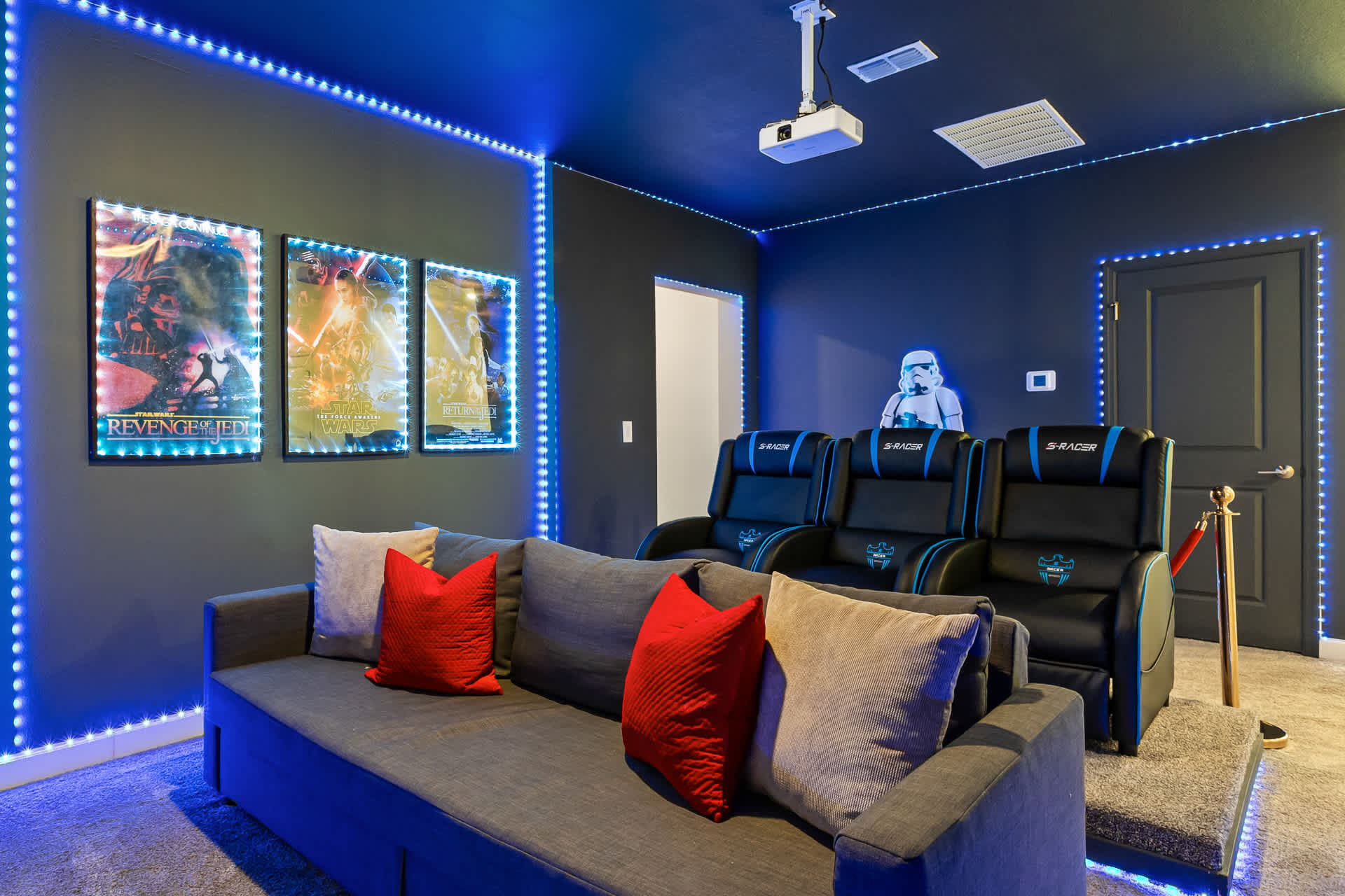 A Star Wars-themed media room with a gray sofa, two black reclining theater seats, and a projector mounted on the ceiling, all illuminated by blue LED strip lights along the walls and ceiling.