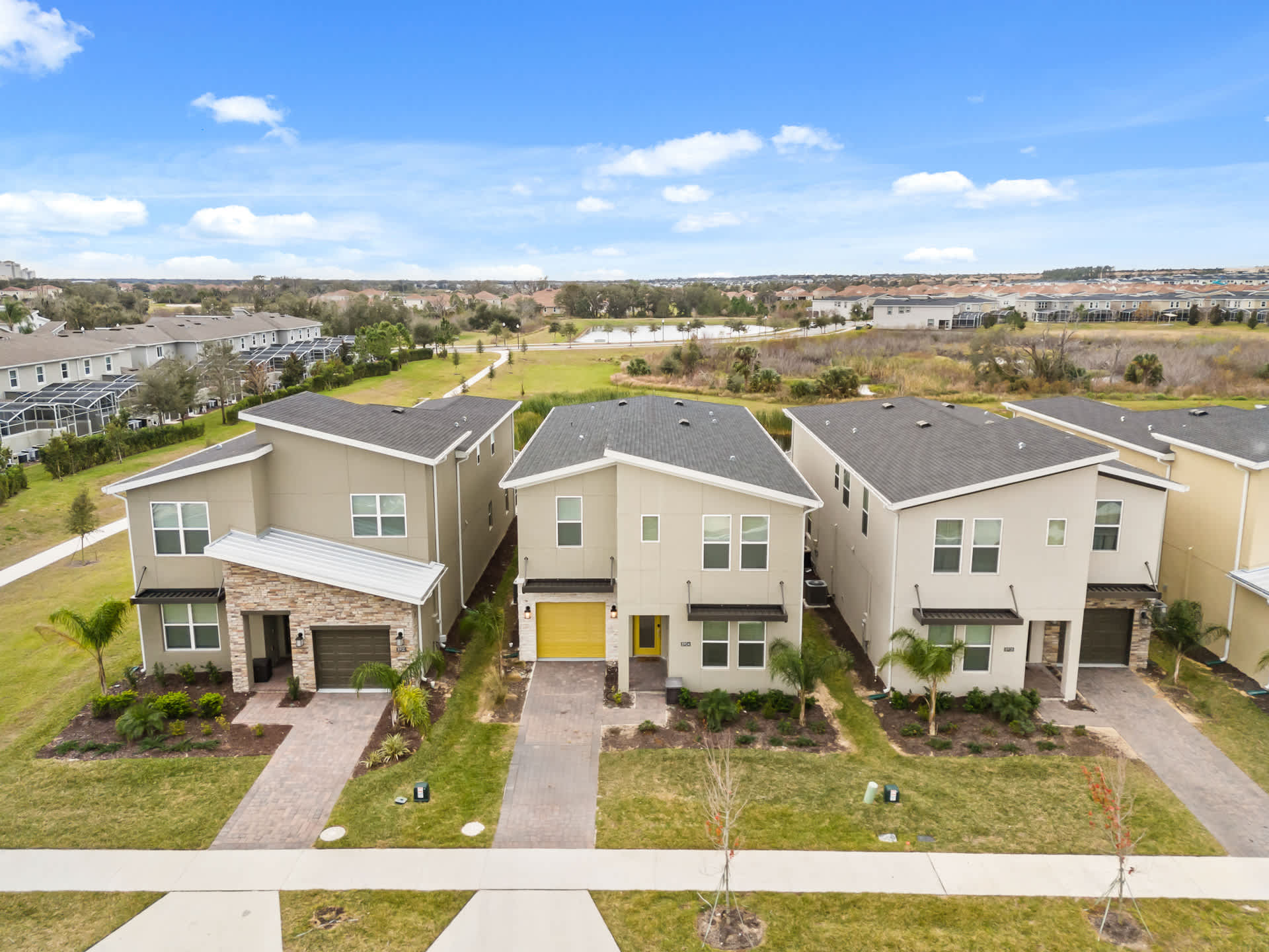 An aerial view showcasing a row of modern two-story vacation homes with light-colored exteriors and dark roofs, each with a driveway and garage. Lush green lawns and landscaping surround the properties, with a pond and open fields in the background under a blue sky.
