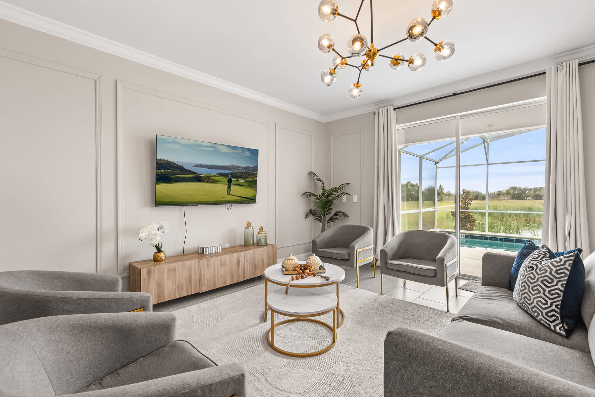 A bright and modern living room with light gray walls, a gray sofa, two gray armchairs, a round marble-top coffee table, and a large TV, offering views of a pool and golf course through a sliding glass door.