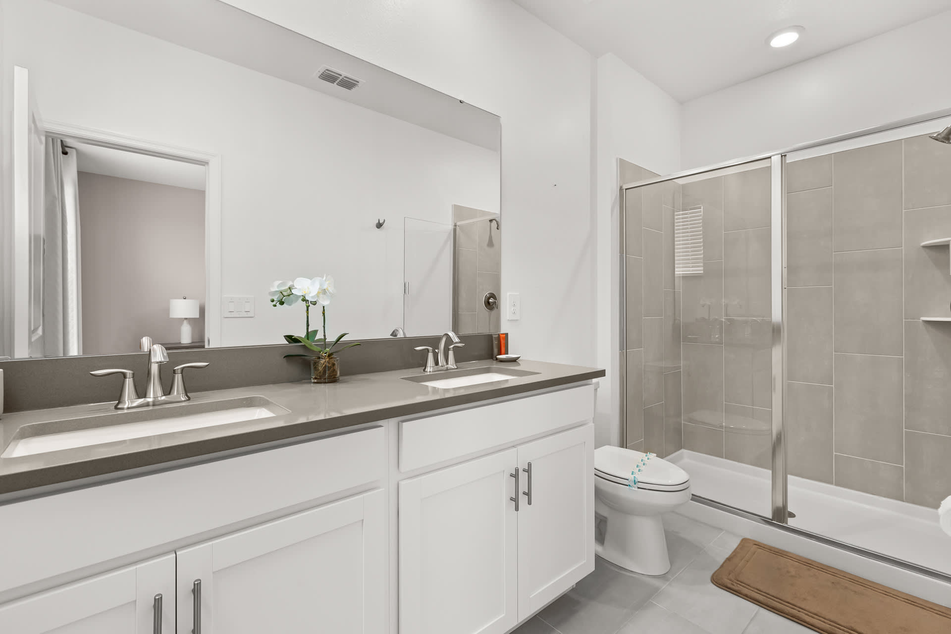 A modern bathroom with a double vanity featuring white cabinets, grey countertops, and rectangular sinks with chrome faucets. A large mirror spans the wall above, and a glass-enclosed walk-in shower with light grey tiled walls is visible on the right.