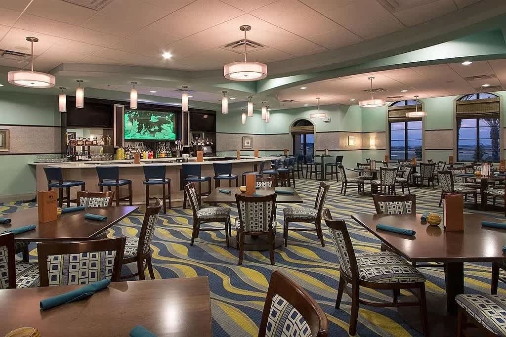 Spacious dining area with multiple wooden tables and chairs featuring patterned upholstery, set on a distinctive blue and yellow wavy carpet. A long bar with blue stools and a large TV screen is visible in the background, under modern pendant lighting.