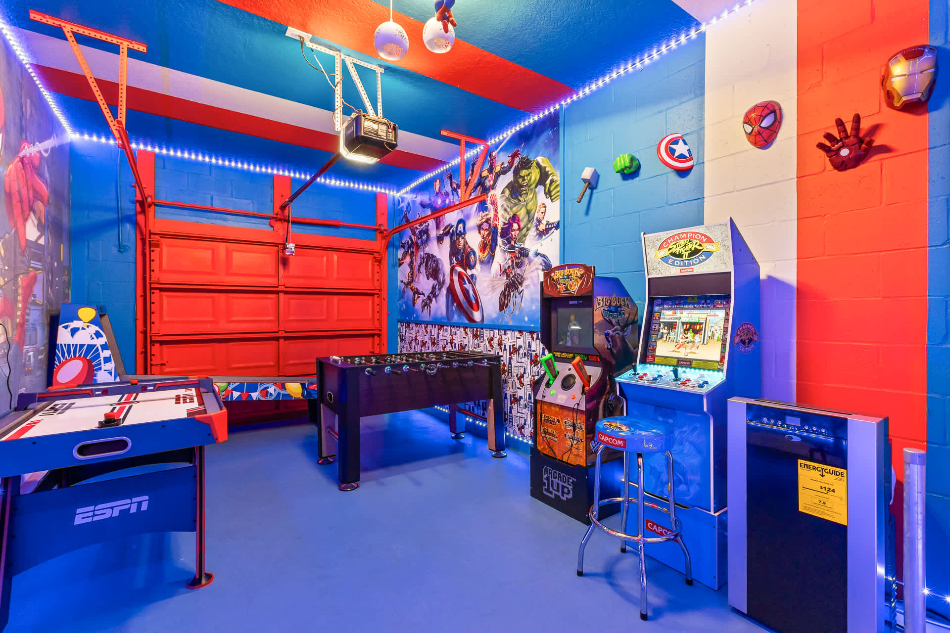 A vibrant, Marvel-themed game room with blue and red walls, featuring an air hockey table, foosball, and two arcade machines, all illuminated by blue LED strip lights. A red garage door is visible in the background.