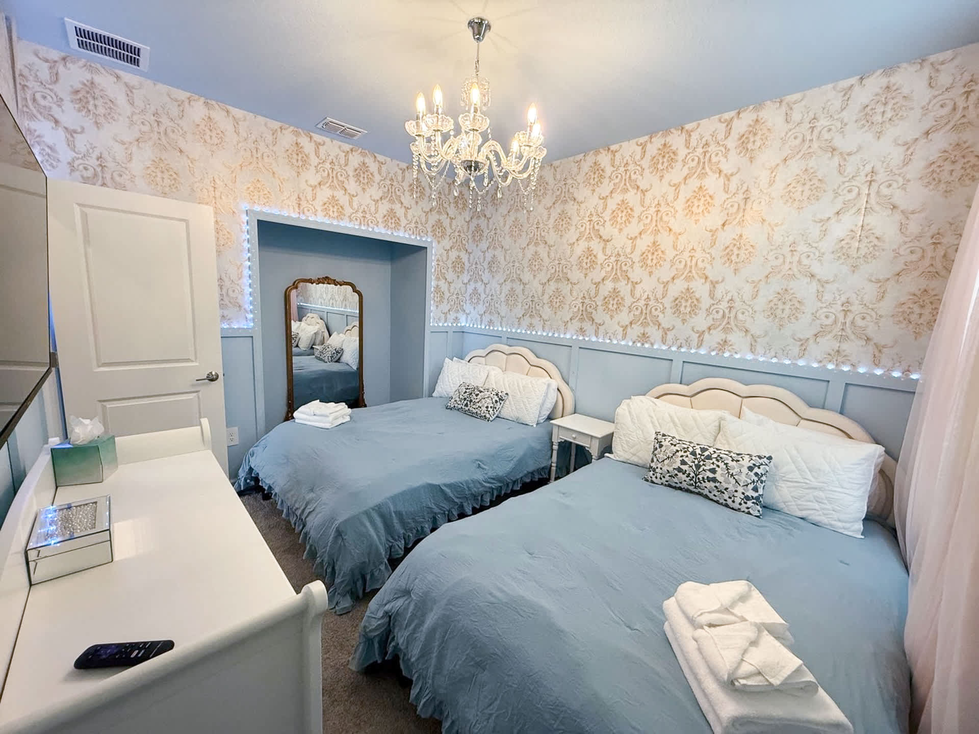 Another angle of the princess-themed bedroom, showcasing two twin beds with light blue bedding and white headboards, and a white dresser with a mirror. The room features ornate gold and white wallpaper, light blue wainscoting, and a crystal chandelier.