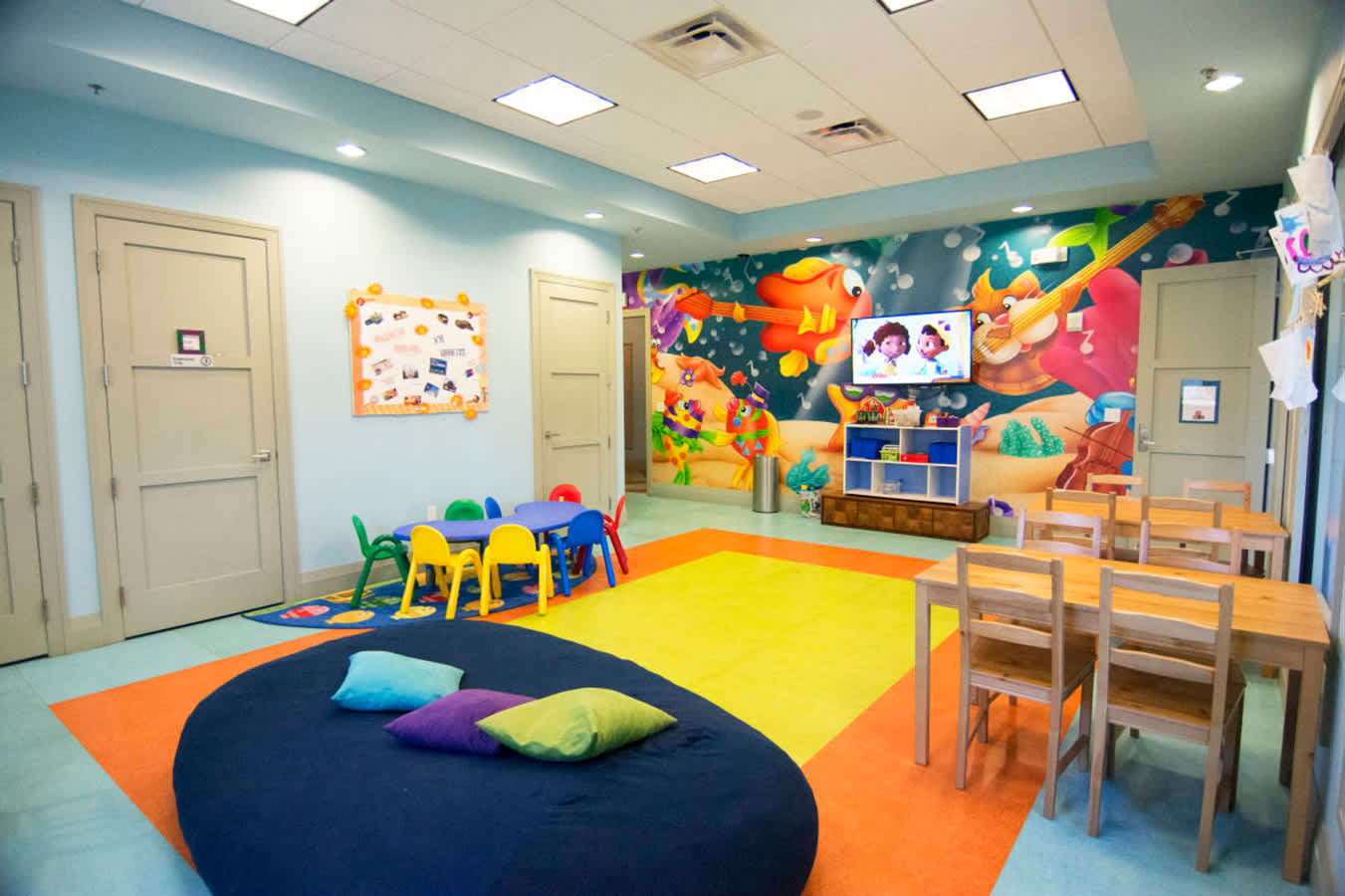 Colorful kids' play room with vibrant wall murals depicting musical instruments and underwater scenes, featuring small tables and chairs, a large beanbag, and a wall-mounted TV. The floor is covered with multi-colored carpet tiles.
