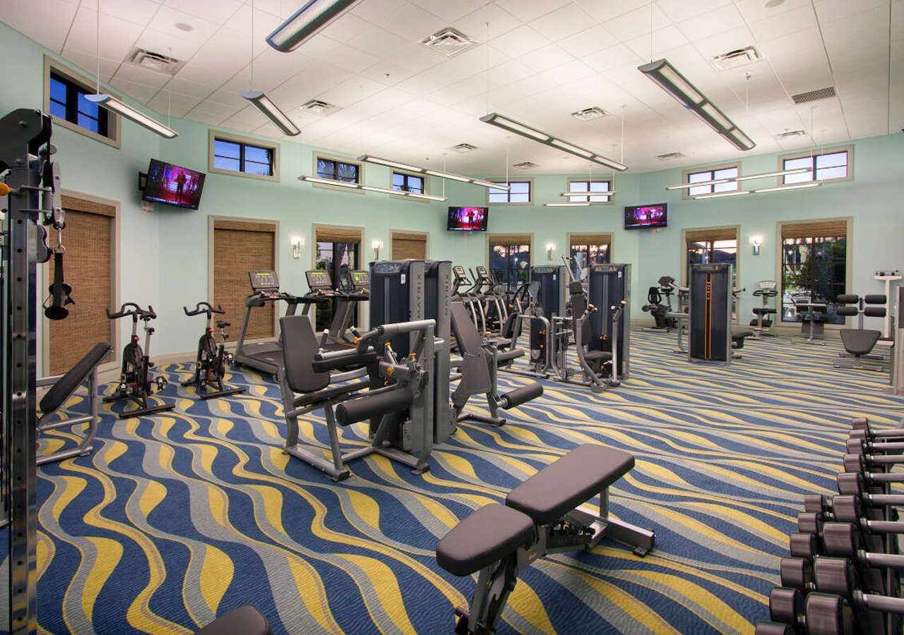 Well-equipped fitness center featuring various exercise machines including treadmills, ellipticals, and weight machines, arranged on a vibrant blue and yellow wavy carpet. Multiple wall-mounted TVs and large windows with blinds are visible.