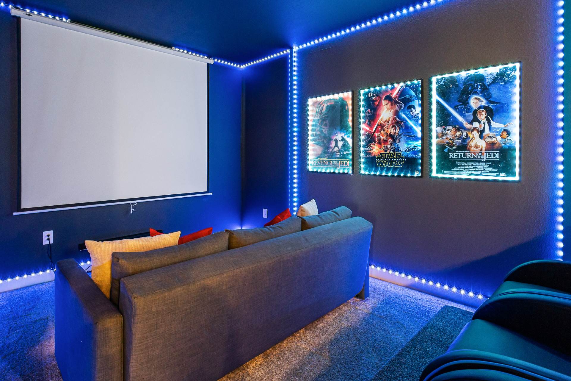 A dark, cozy media room featuring a large projector screen, a comfortable gray sofa with red and beige pillows, and three backlit Star Wars movie posters, all outlined by blue LED strip lighting.