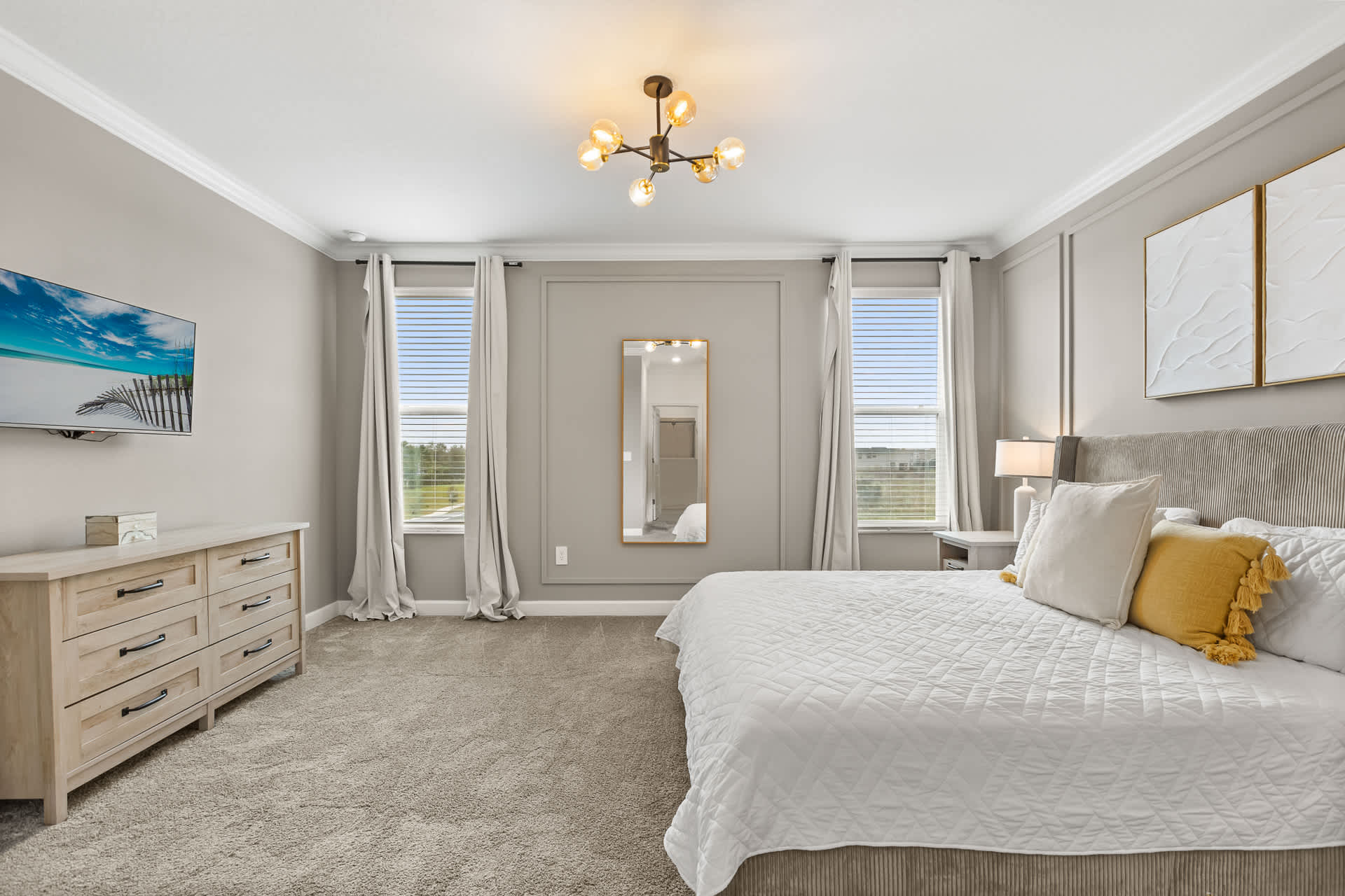 Another view of the spacious bedroom with a queen-sized bed, light gray carpet, and paneled walls, showing a wall-mounted TV above a light wood dresser and two windows with blinds and curtains.