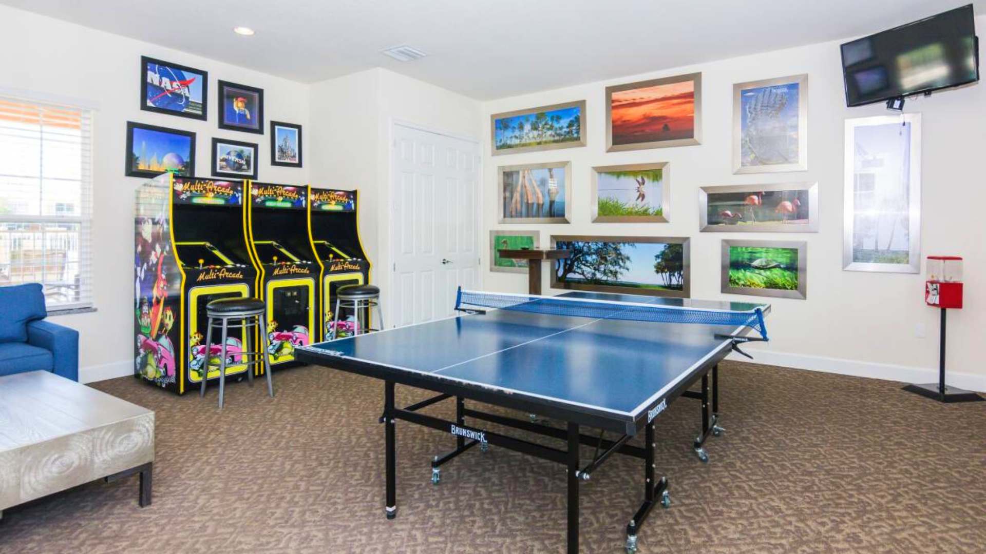 A lively game room featuring a blue ping pong table in the center, two vintage-style arcade machines with stools, and a collection of framed artwork on the walls, all set on a patterned brown carpet.