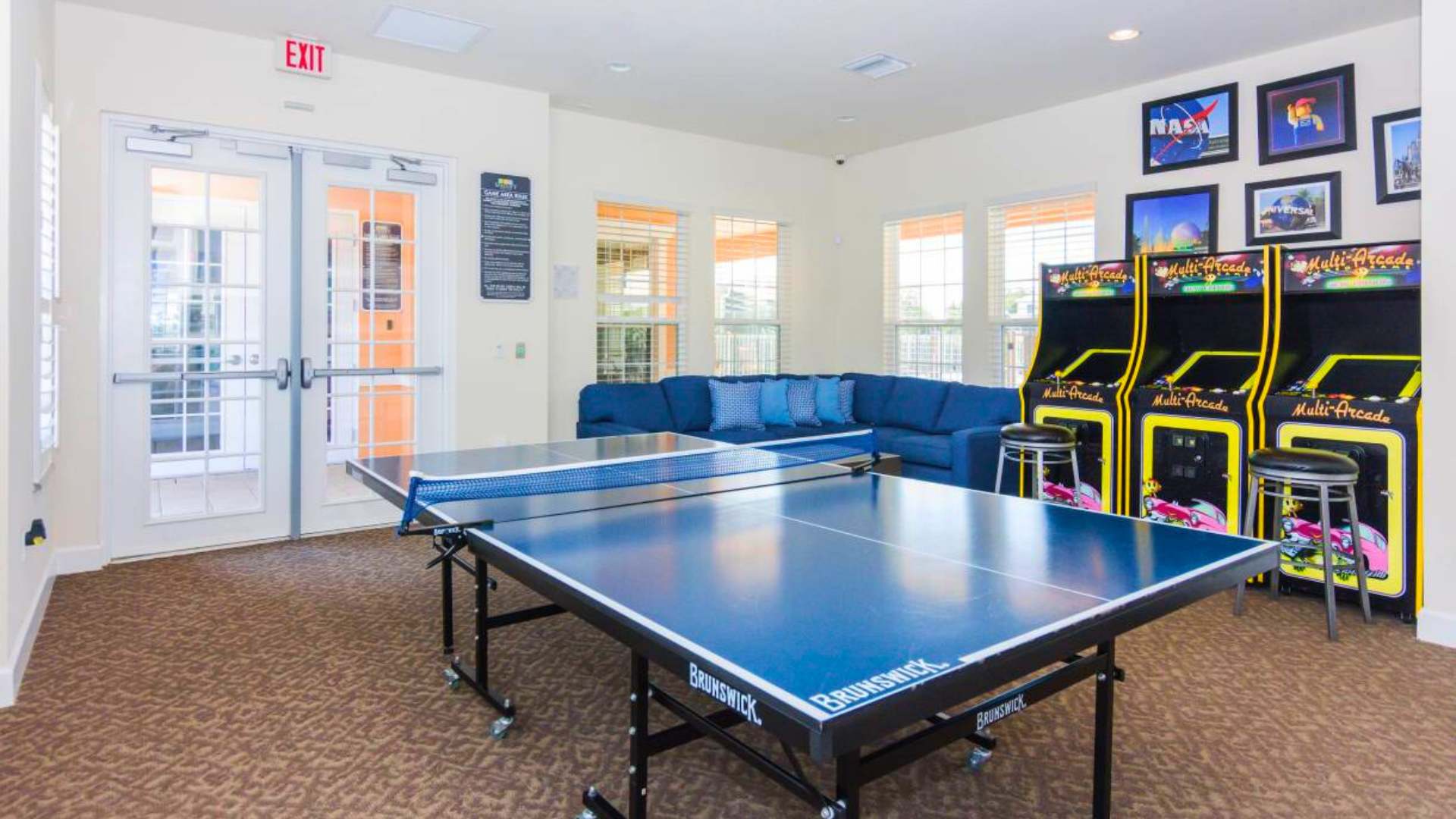 A spacious game room with a blue ping pong table, two arcade machines, and a comfortable blue sectional sofa, featuring large windows and glass exit doors, all on a patterned brown carpet.