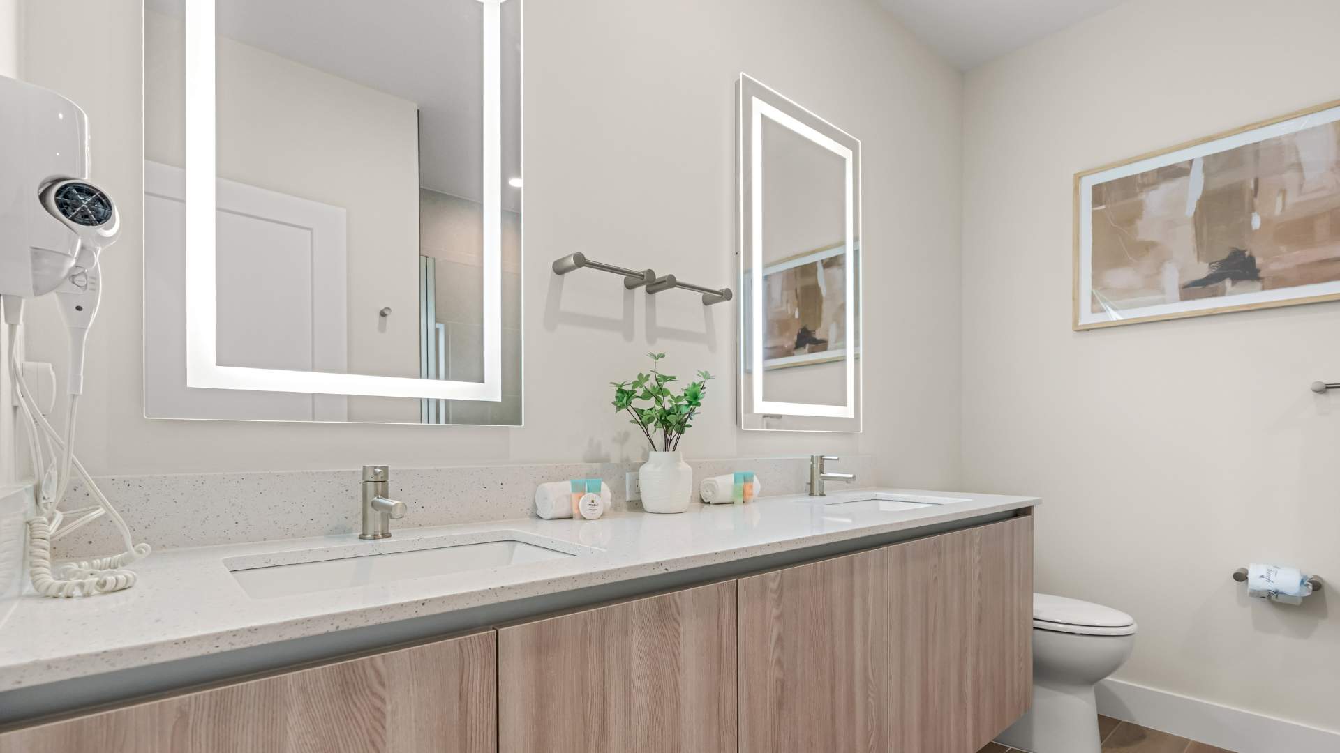 Modern bathroom featuring a double vanity with light-colored quartz countertops, two rectangular sinks, and two large backlit mirrors. A white hairdryer is mounted on the left wall, and a small plant decorates the counter.