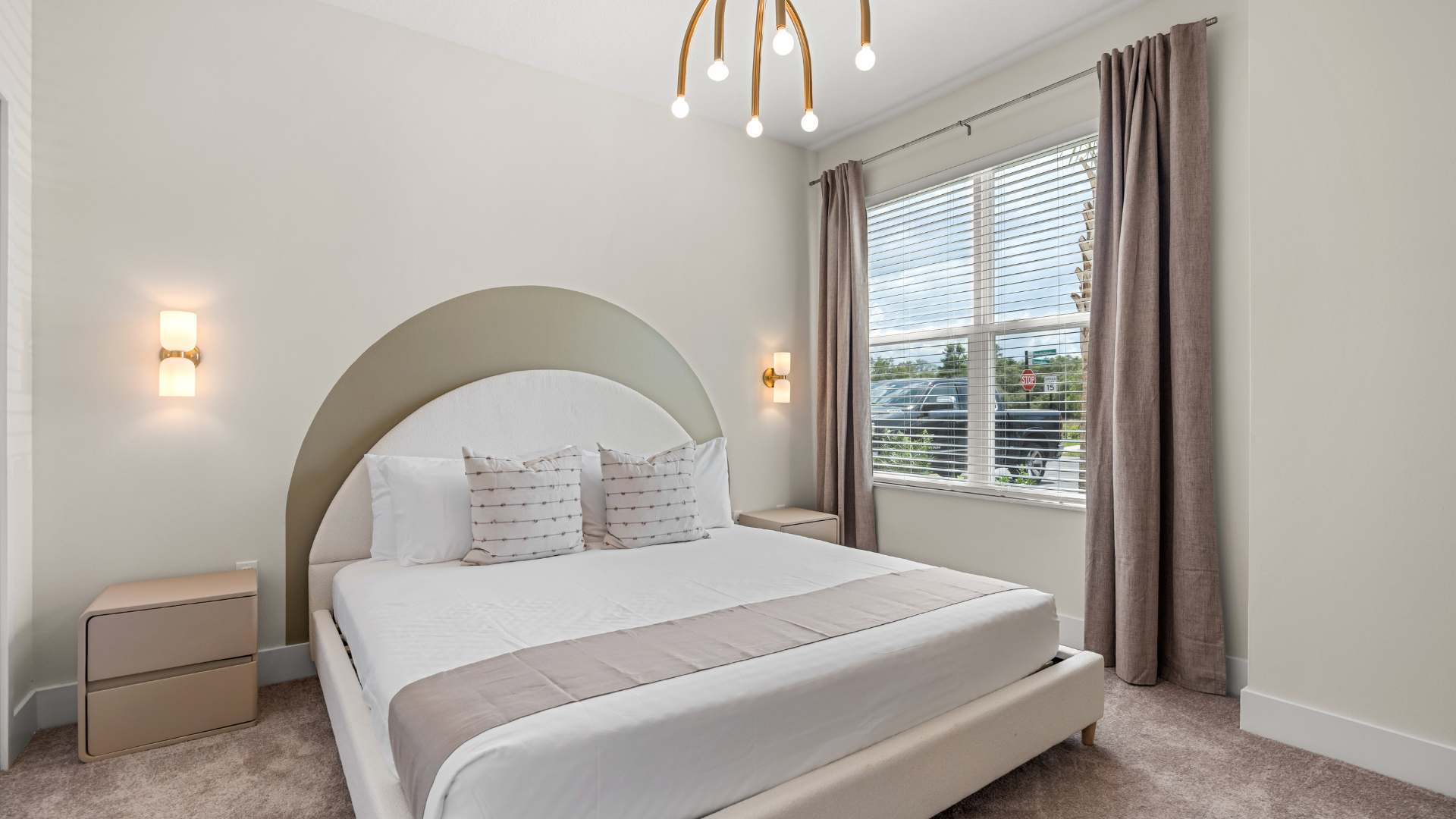 Modern bedroom with a white upholstered bed featuring an arched headboard, flanked by light-colored nightstands and wall sconces. A large window with blinds and curtains provides natural light.
