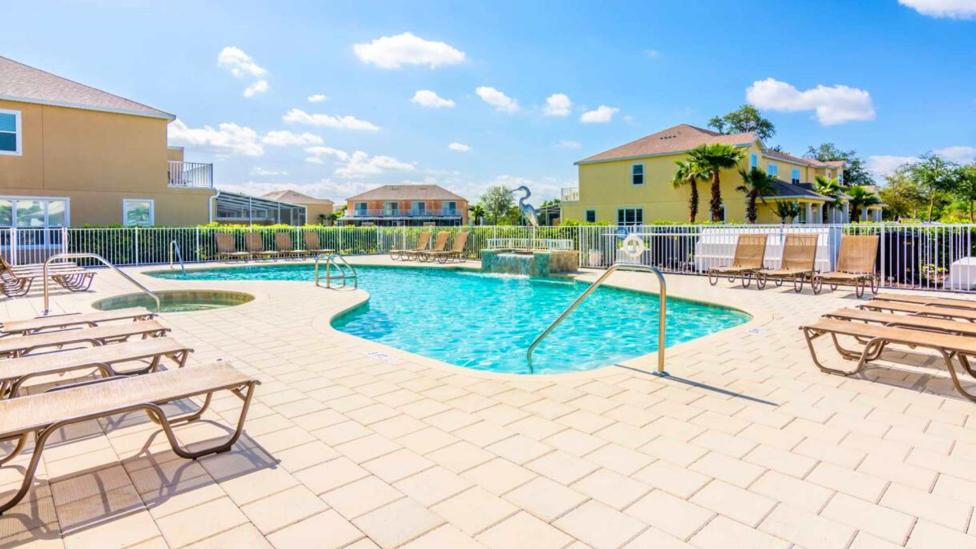 An inviting communal swimming pool with an adjacent hot tub, both featuring clear blue water, surrounded by a spacious light-colored paver deck with numerous lounge chairs and a white fence, with residential buildings in the background.