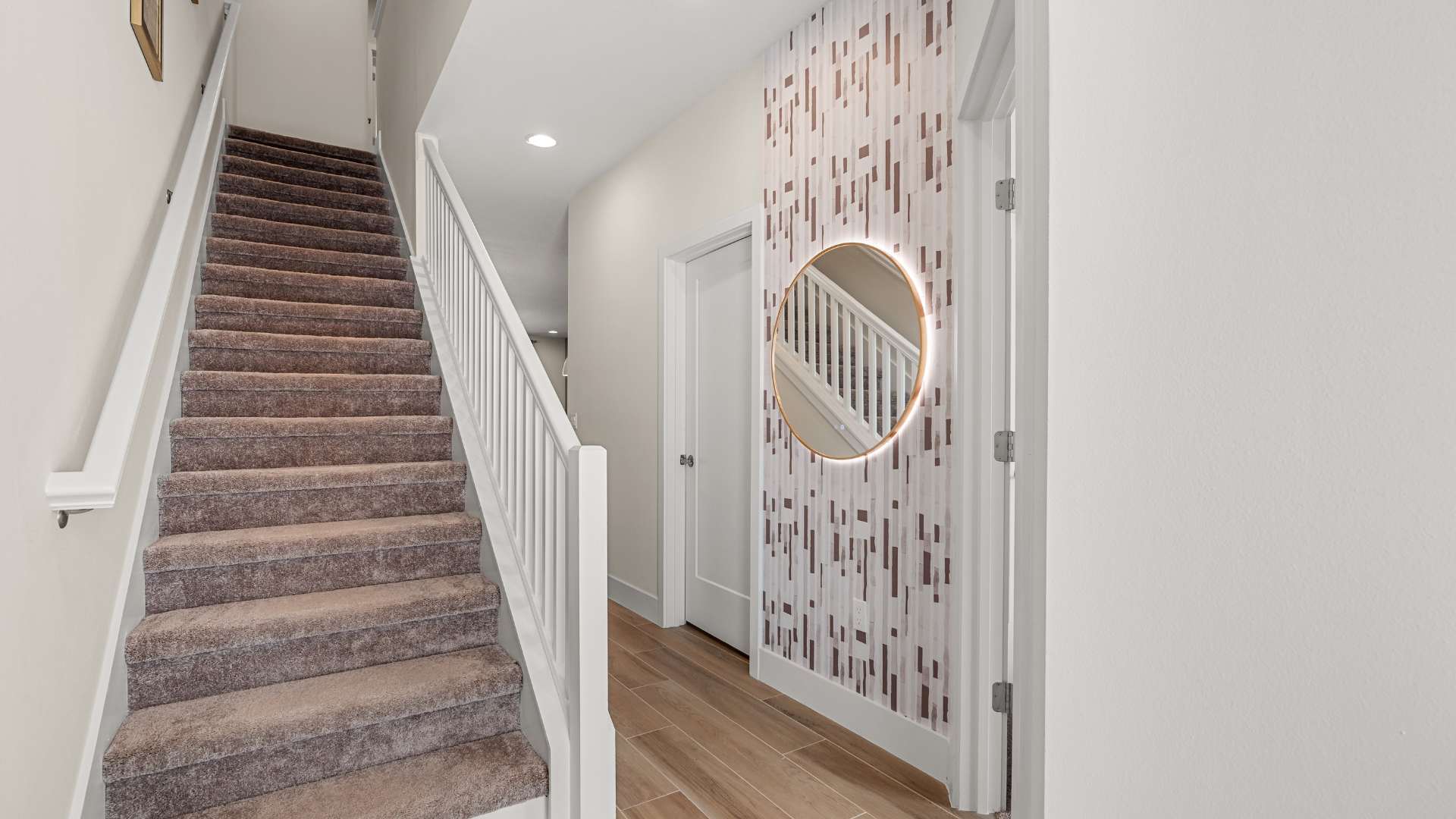 Carpeted staircase with white railings leading upstairs, adjacent to a hallway with a decorative vertical wood panel wall and a round gold-framed mirror. Light wood-look flooring is visible.