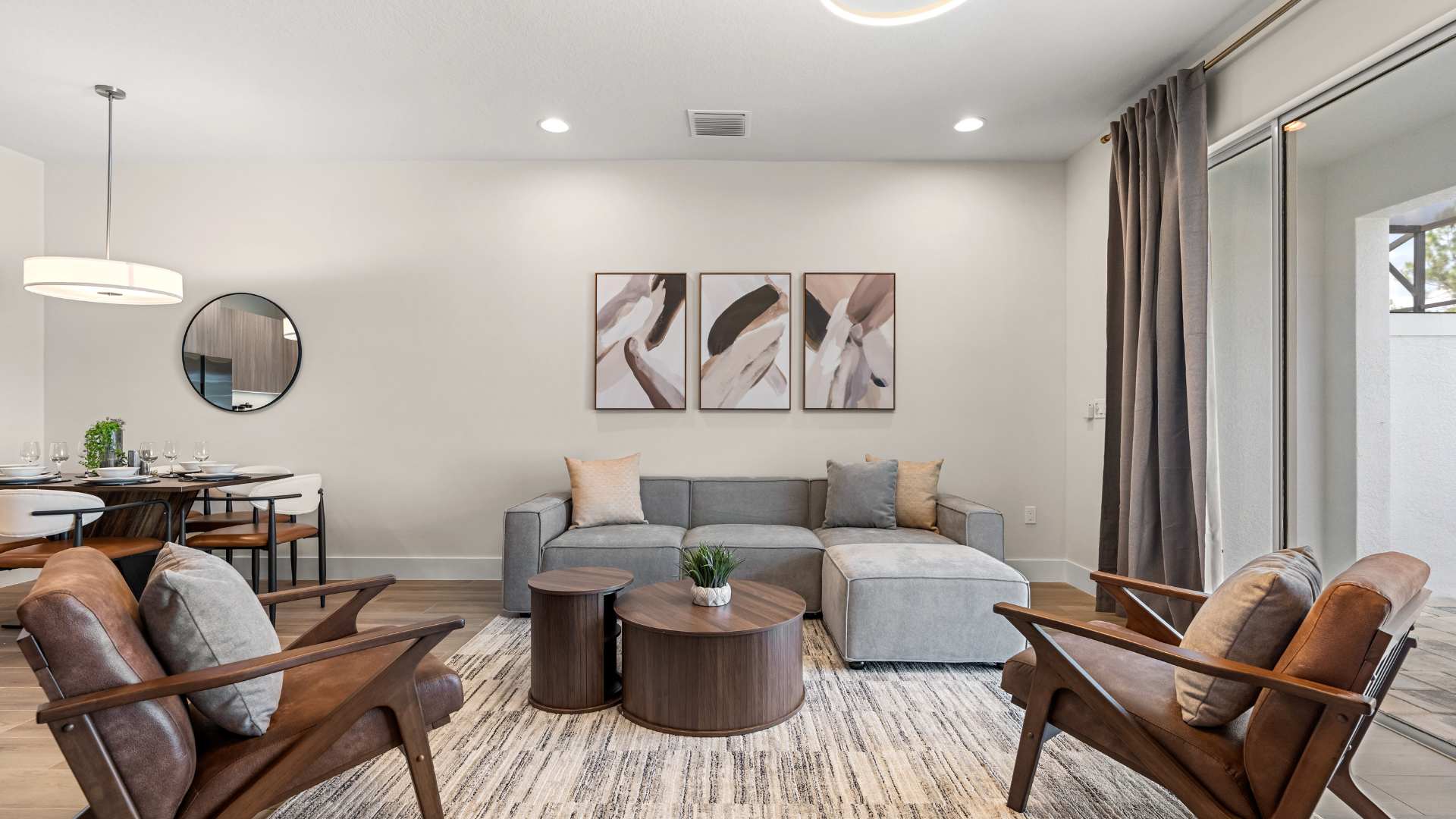 Cozy living room featuring a gray sectional sofa with throw pillows, two brown leather armchairs, and a striped area rug on light wood floors. Three abstract art pieces adorn the wall above the sofa, and a modern chandelier hangs from the ceiling.