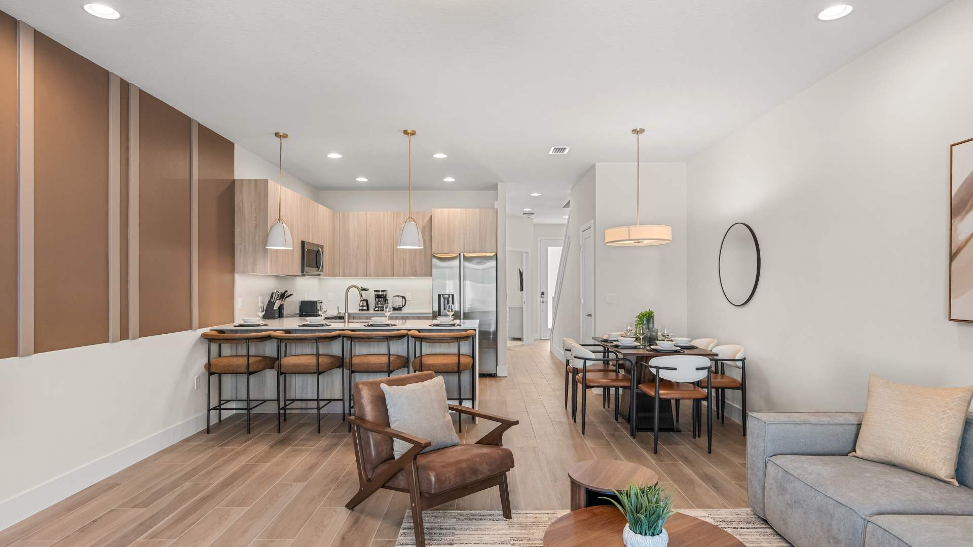 Wide view of the open-concept living space, showcasing the kitchen with a large island, the dining area with a wooden table, and the living room with a gray sectional sofa and brown leather armchairs. Light wood floors connect all areas.
