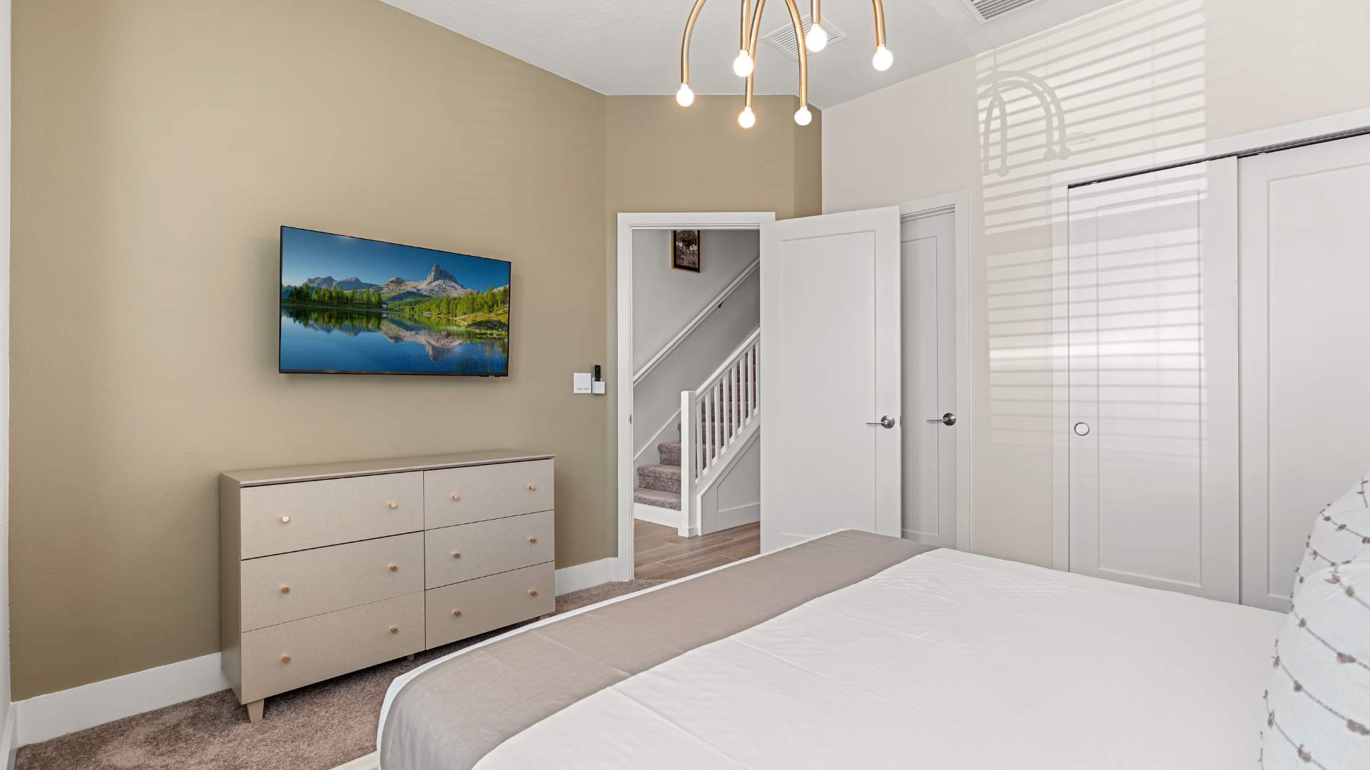 Bedroom featuring a light-colored dresser with gold hardware and a wall-mounted flat-screen TV. An open doorway reveals a staircase, suggesting proximity to common areas.