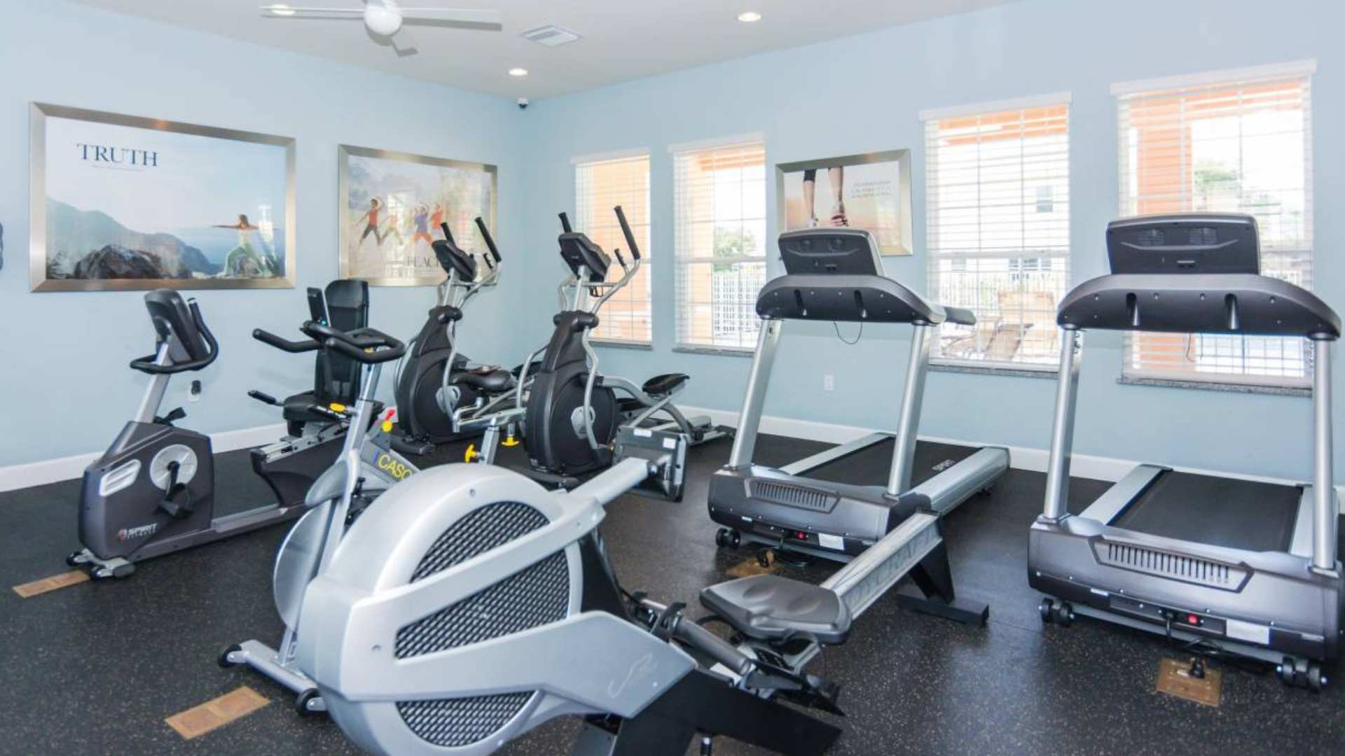 A well-equipped fitness room featuring multiple treadmills, elliptical machines, and a stationary bike, set on a black rubber floor with light blue walls and large windows, providing a bright workout space.