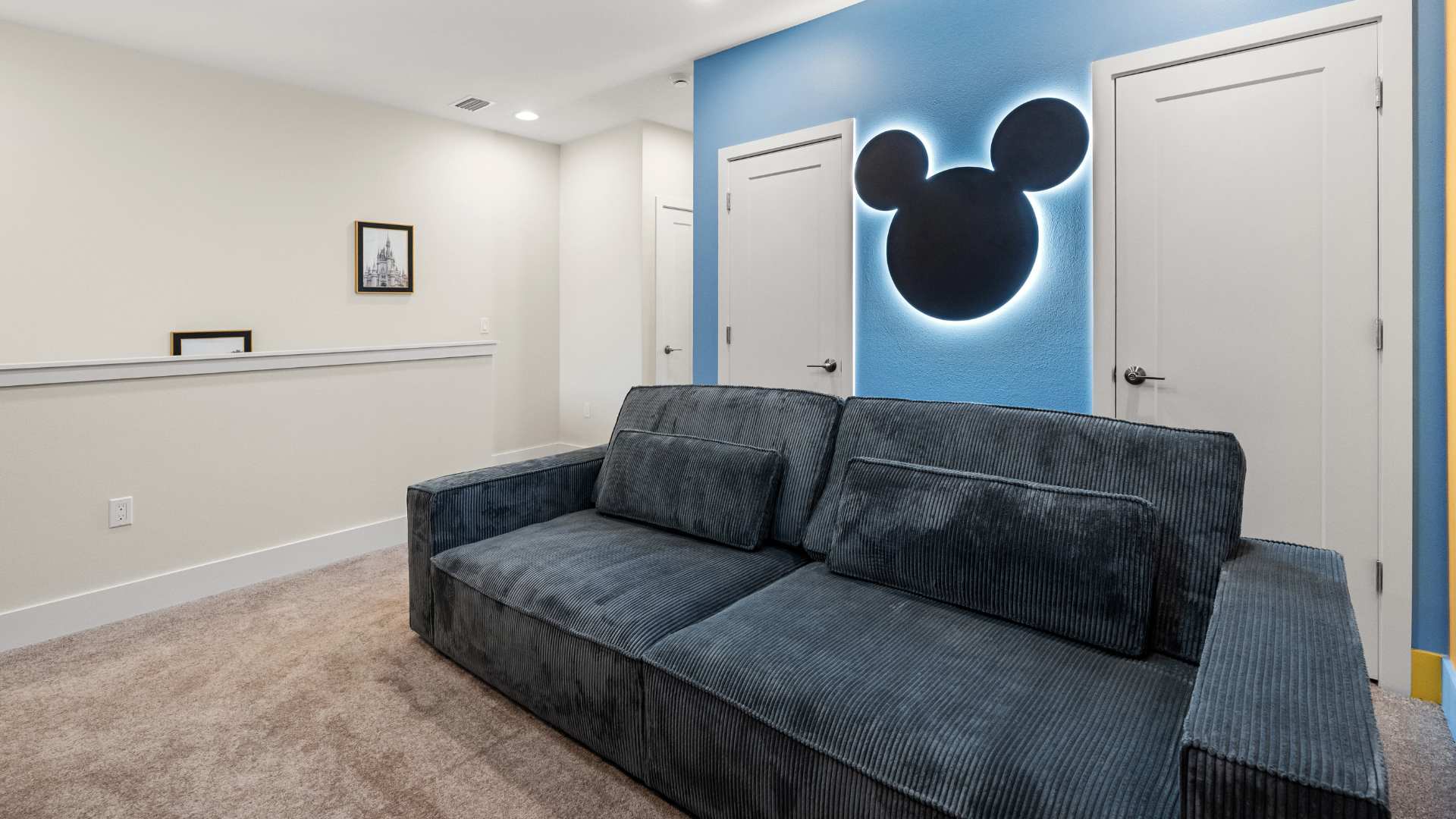Cozy media room featuring a large dark gray sofa and a vibrant blue accent wall with a backlit Mickey Mouse silhouette decoration. The room has carpeted flooring.